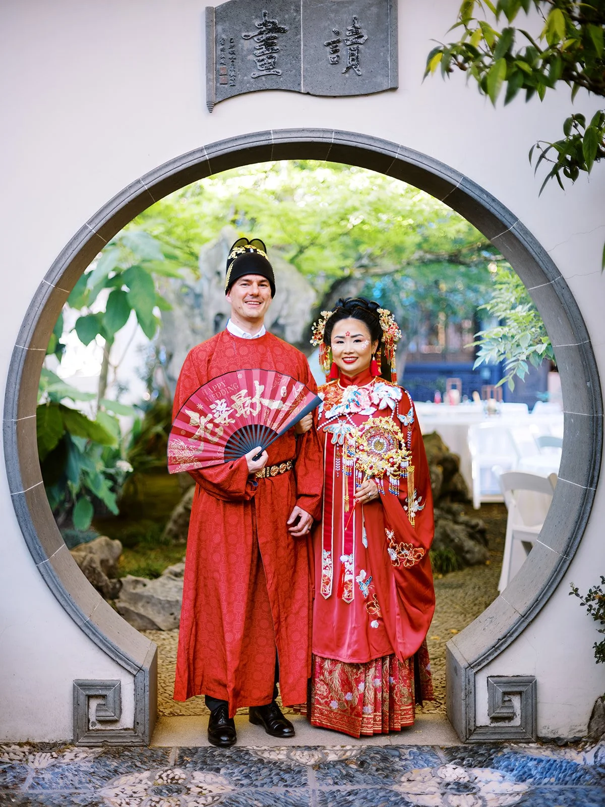 Bride and groom standing together inside a circular moon gate, smiling at the camera with the garden reception tables visible in the background