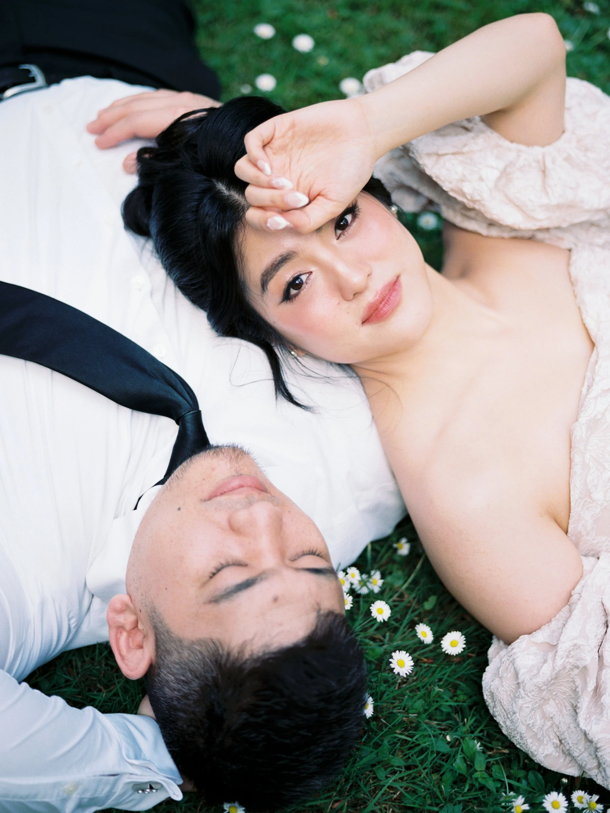 Wedding couple lying together in the grass surrounded by small white flowers.