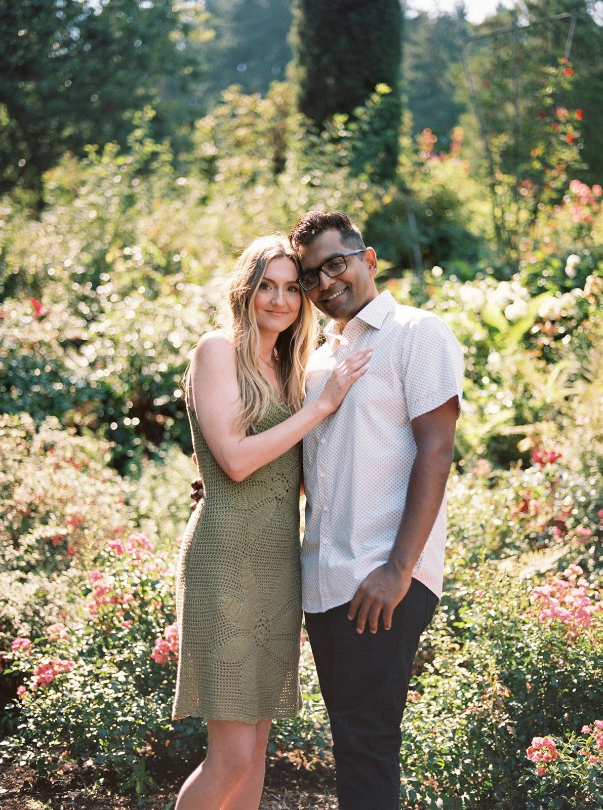 A couple leaning together and smiling in a sun-drenched rose garden filled with soft pink blooms.