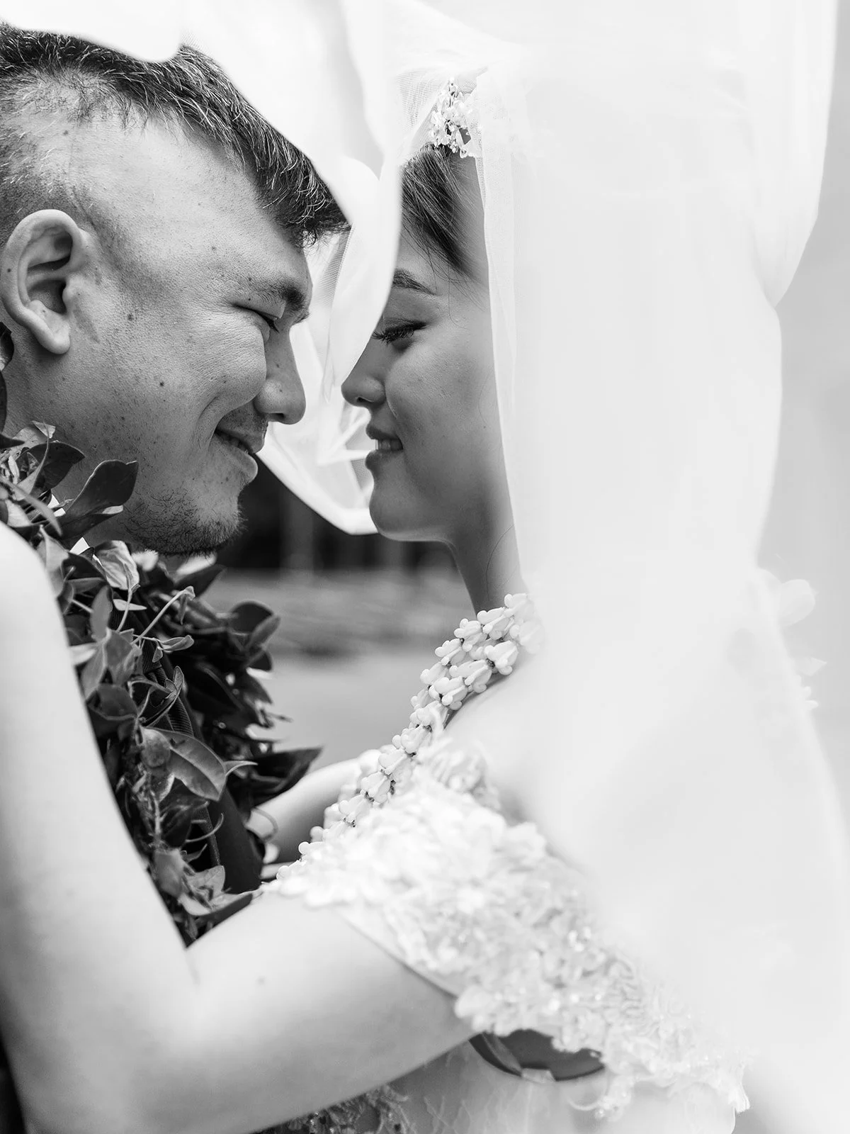 Black and white close-up of the bride and groom nearly nose-to-nose under her veil, both wearing Hawaiian lei — his a green maile lei, hers a white pikake lei