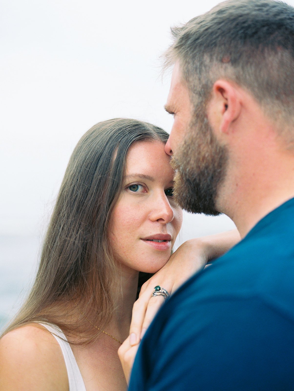 Close portrait of couple embracing on the beach, woman resting her hand on man’s shoulder.