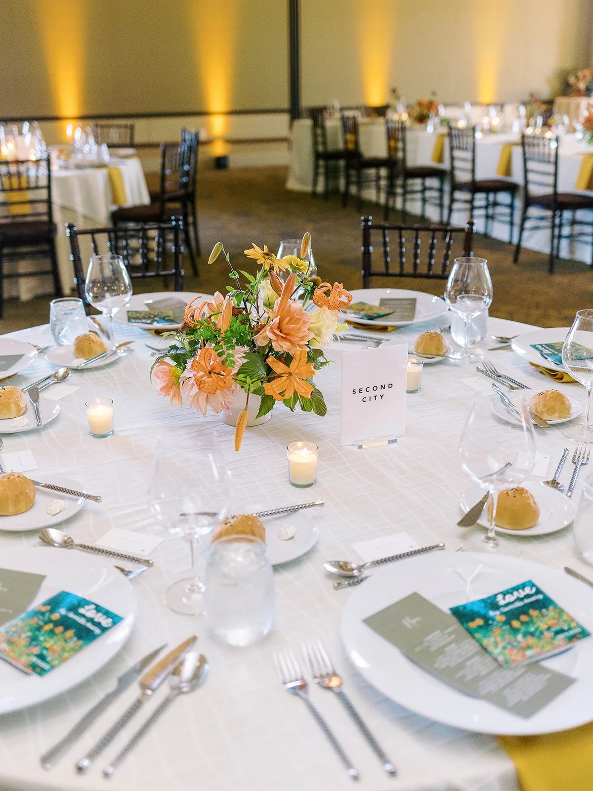 Round reception table set with white linens, yellow napkins, orange and peach floral centerpiece, candles, menus, and a table card reading "Second City," with the full ballroom visible in the background