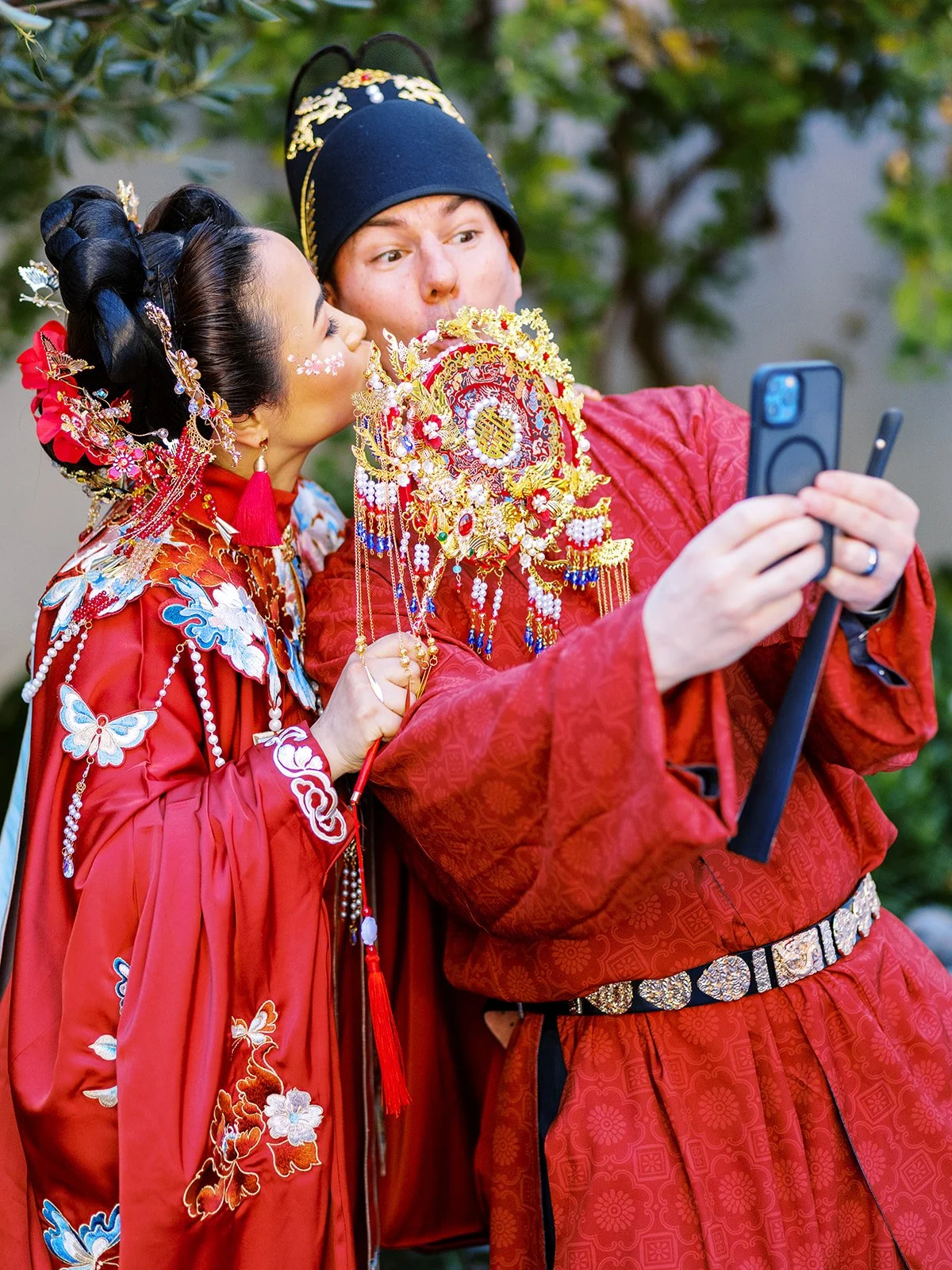 Bride kissing the groom on the cheek while he takes a selfie, both in full red traditional Chinese wedding attire with the bride's elaborate gold bridal bouquet between them
