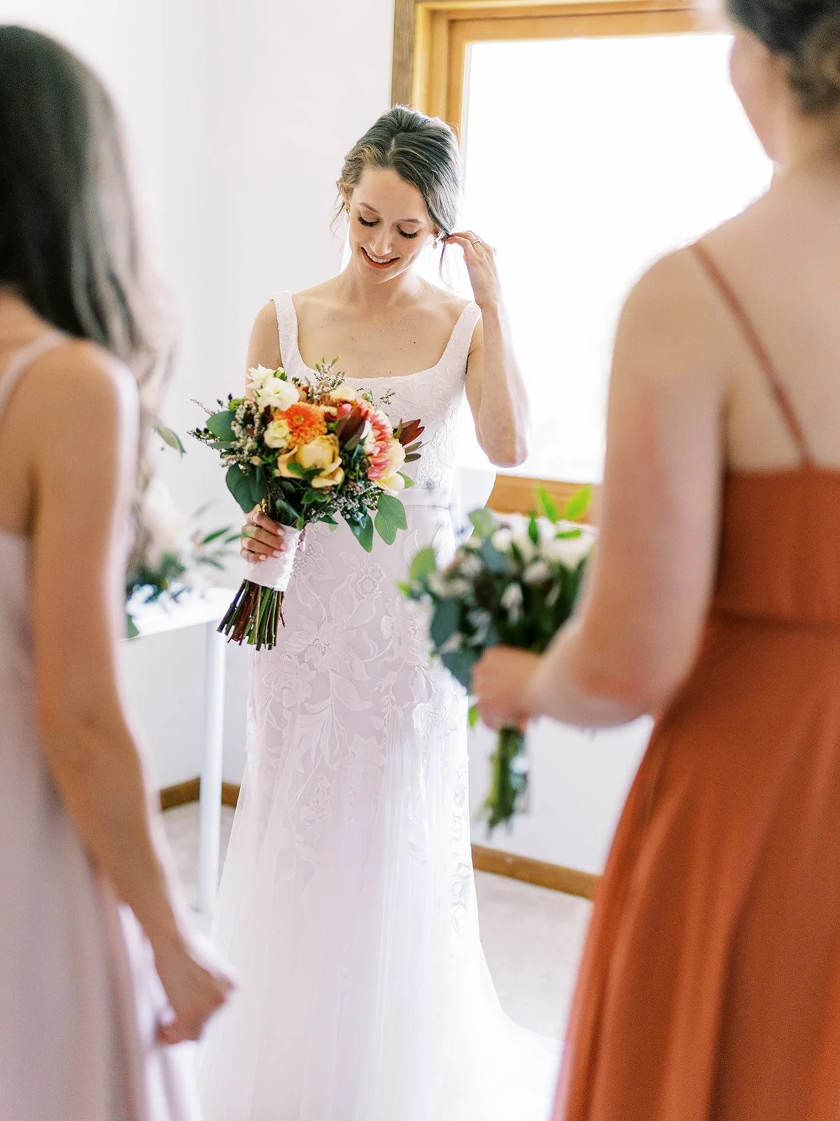 Bride in lace wedding dress holds a colorful bouquet while bridesmaids stand nearby in a bright room.
