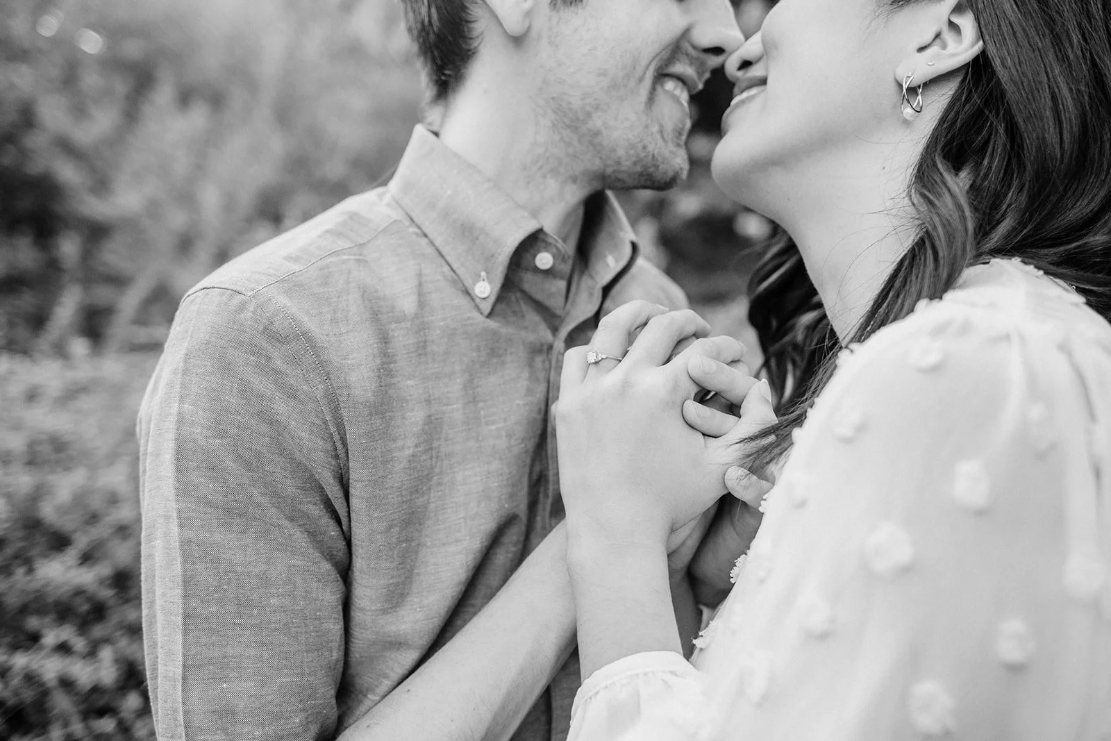 Close-up of couple holding hands, engagement ring visible, faces softly out of focus in outdoor setting