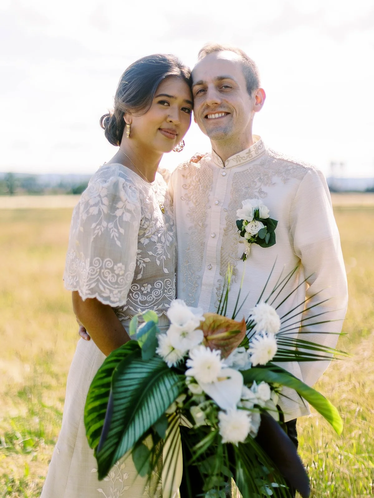 Bride and groom posing together in a golden field, holding a tropical white bouquet with lush greenery
