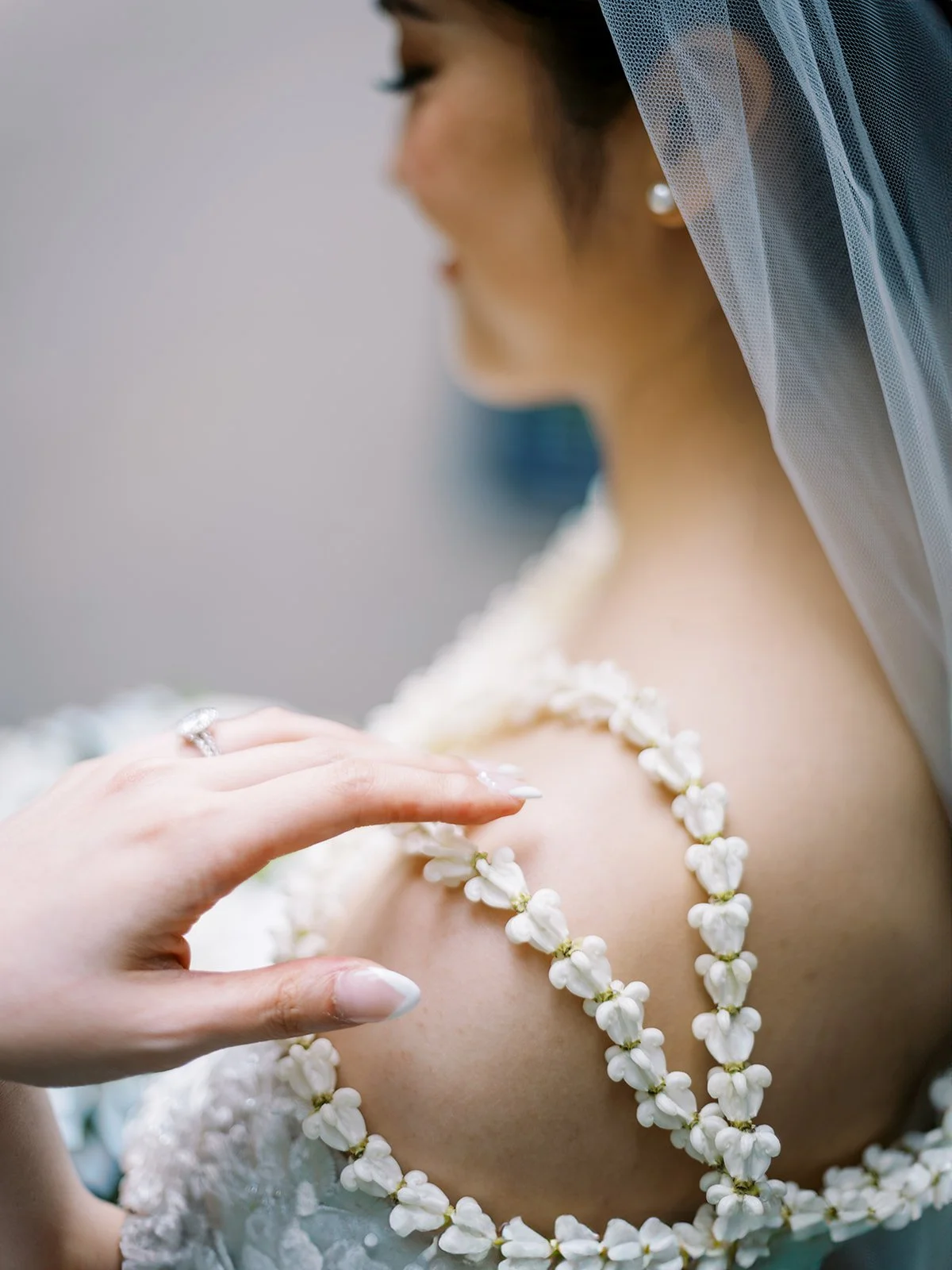 Close-up detail of the bride's white pikake lei draped over her bare shoulder, her engagement ring and french-tipped nails visible as she adjusts it