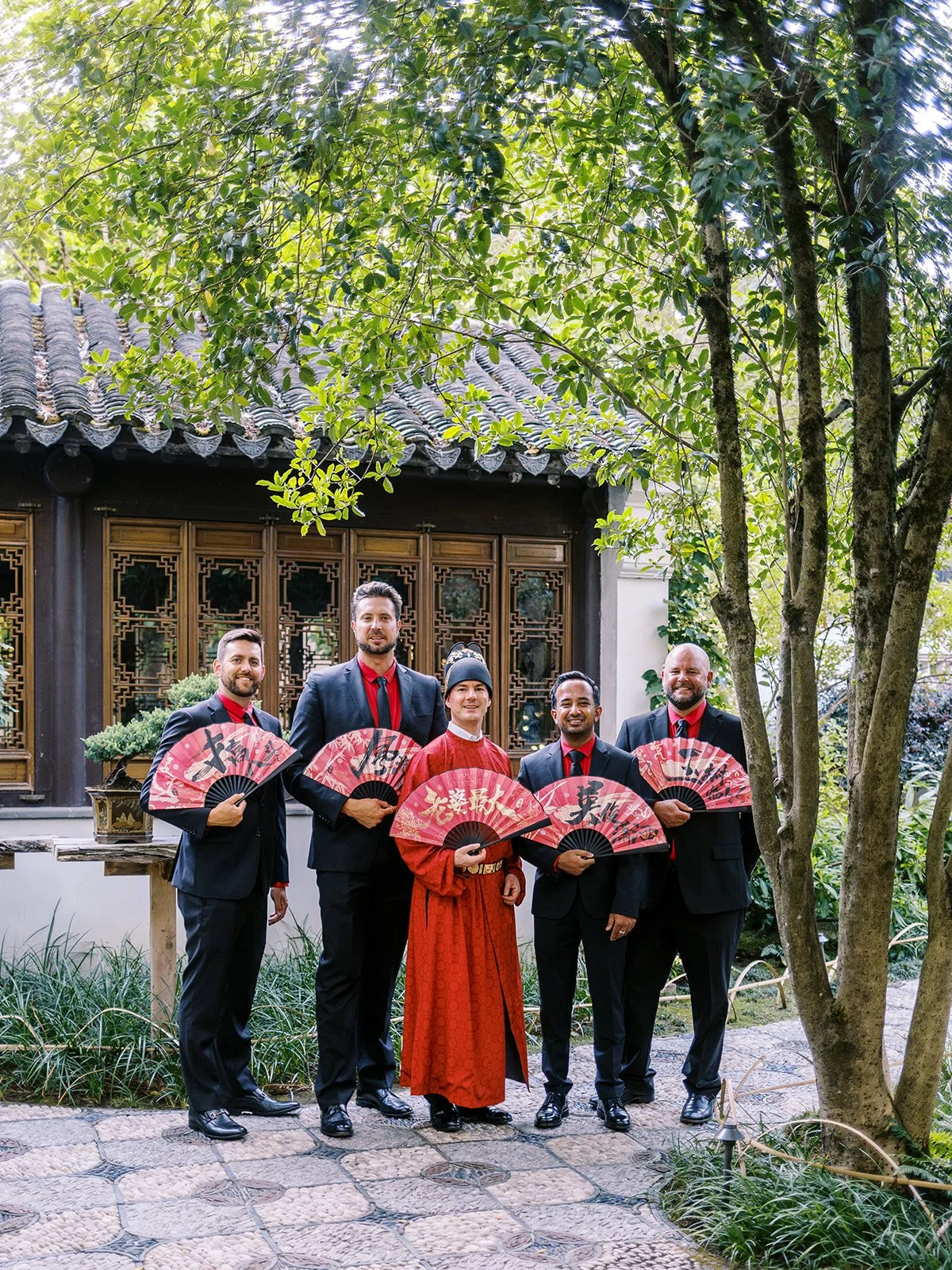 Groom in red traditional Chinese wedding robe and ceremonial hat posing with four groomsmen in black suits and red ties, all holding red fans in front of a garden pavilion