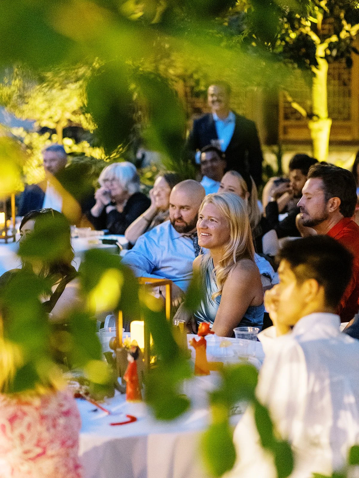 Candlelit evening reception; guests seated at white-clothed tables look on attentively, framed by glowing garden greenery and warm uplighting.