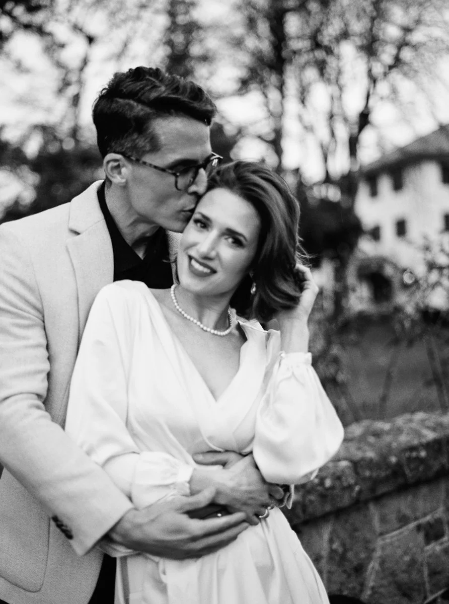 Black and white close-up of couple, man kissing woman’s temple as she smiles, wearing pearl necklace and soft waves