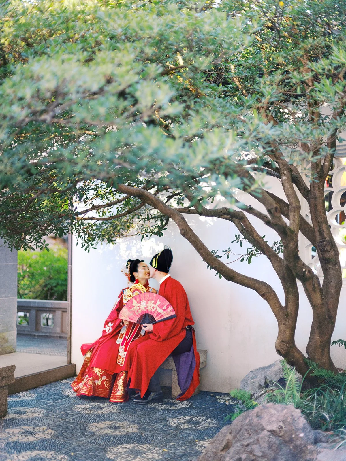 Bride and groom in matching red traditional Chinese wedding attire sharing a quiet moment on a stone bench beneath a garden tree at Lan Su