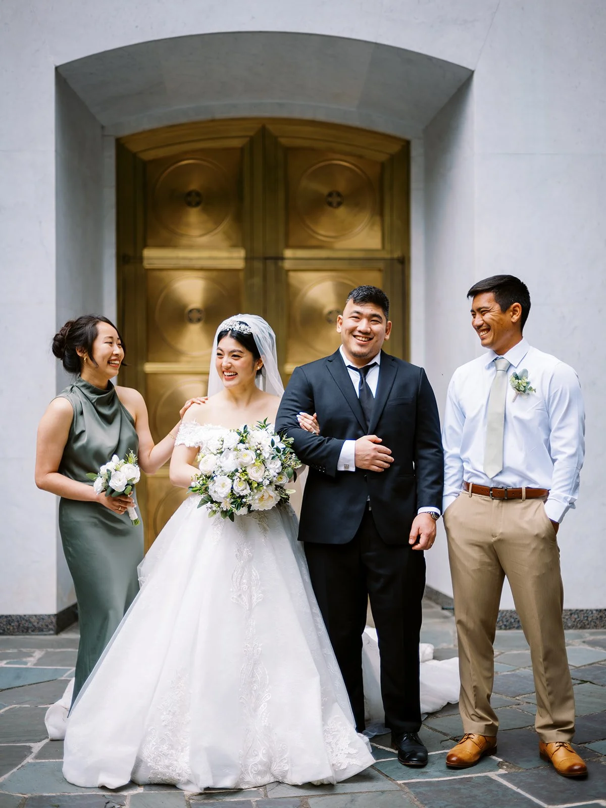 Bride and groom laughing with two friends outside The Grotto's gold doors — a bridesmaid in a sage green dress and a groomsman in a light suit