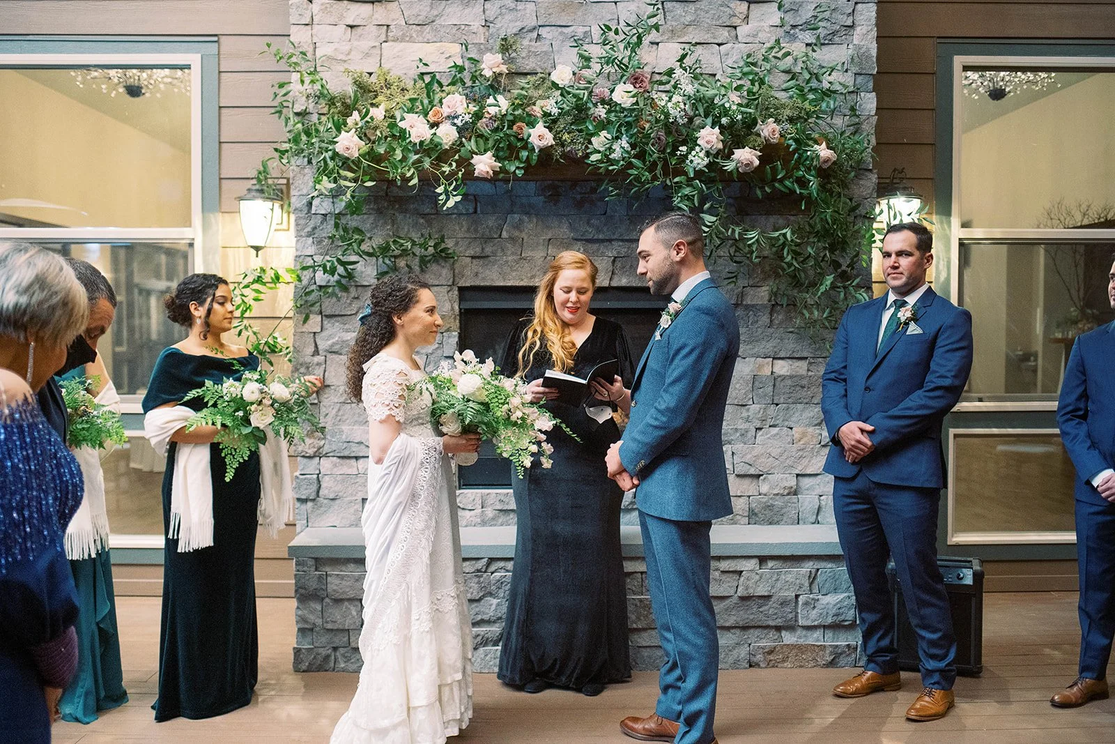 Bride and groom stand with their officiant before a flower-covered stone fireplace during the ceremony.