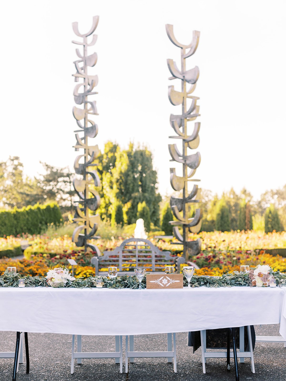 Long outdoor wedding reception table decorated with greenery, flowers, candles and glassware in a colorful garden setting.
