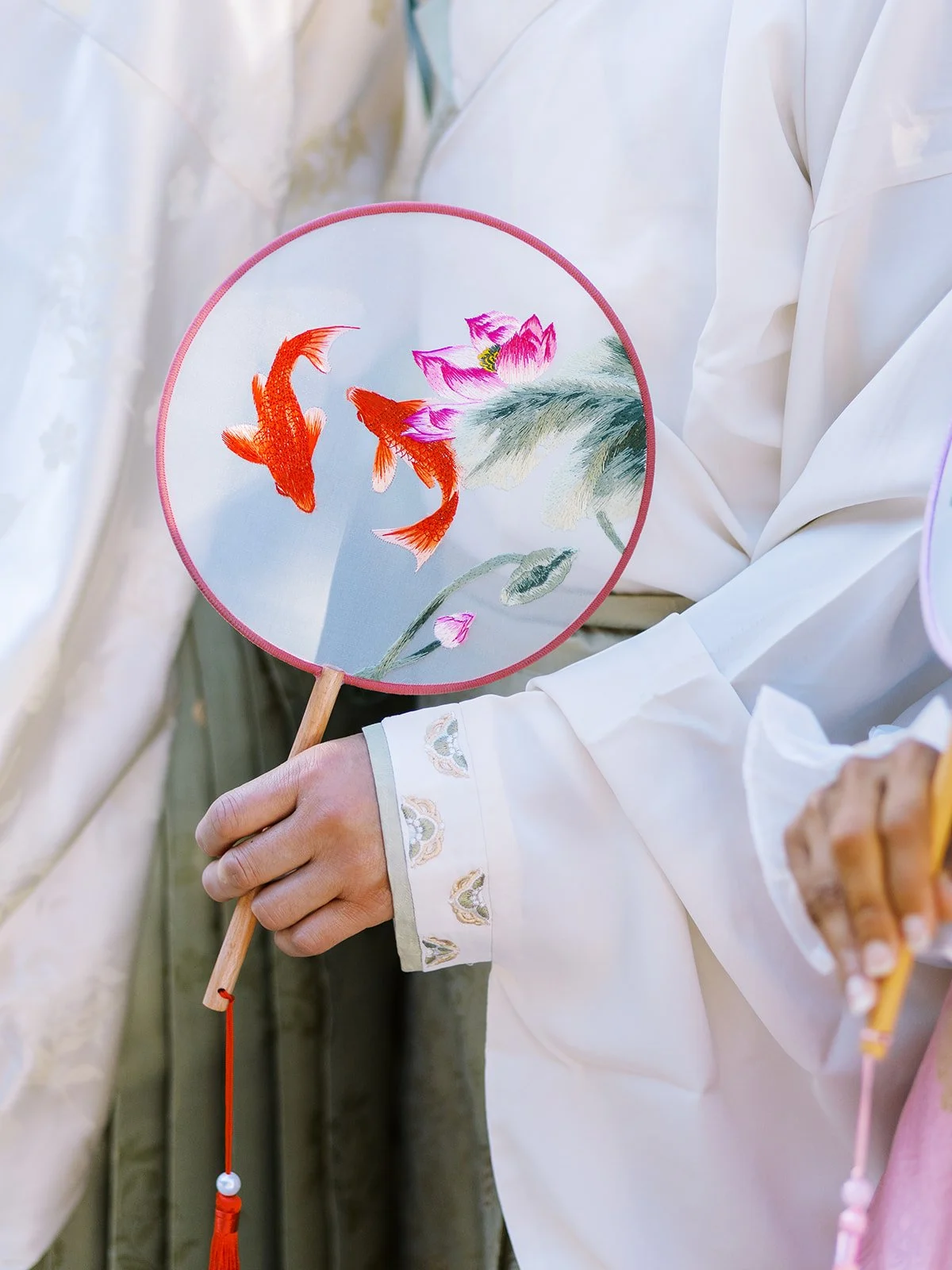 Hand holding a round fan embroidered with goldfish and lotus flowers against soft neutral clothing