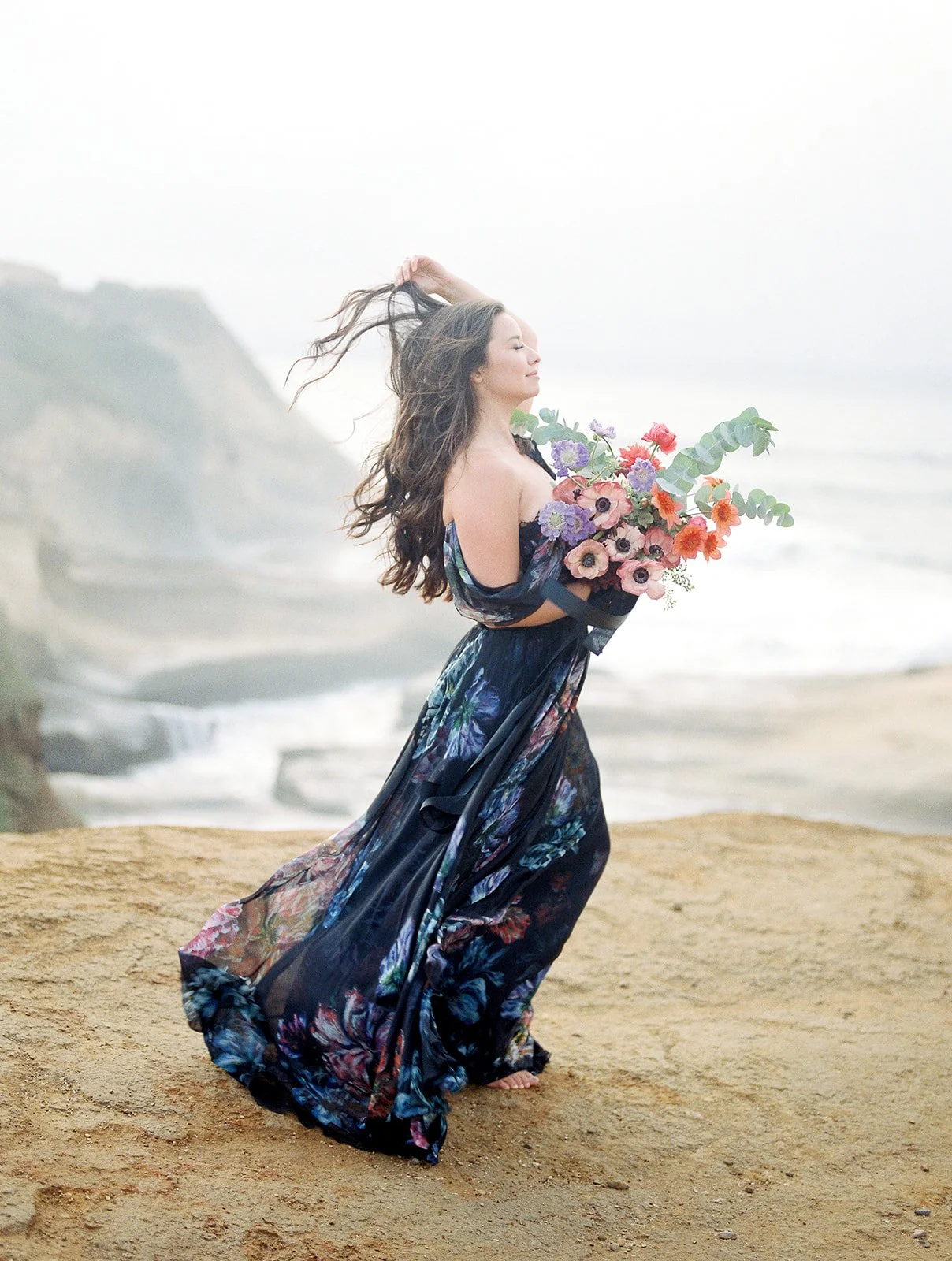 Woman in long floral dress holding bouquet, hair blowing in wind on sandy oceanfront cliff