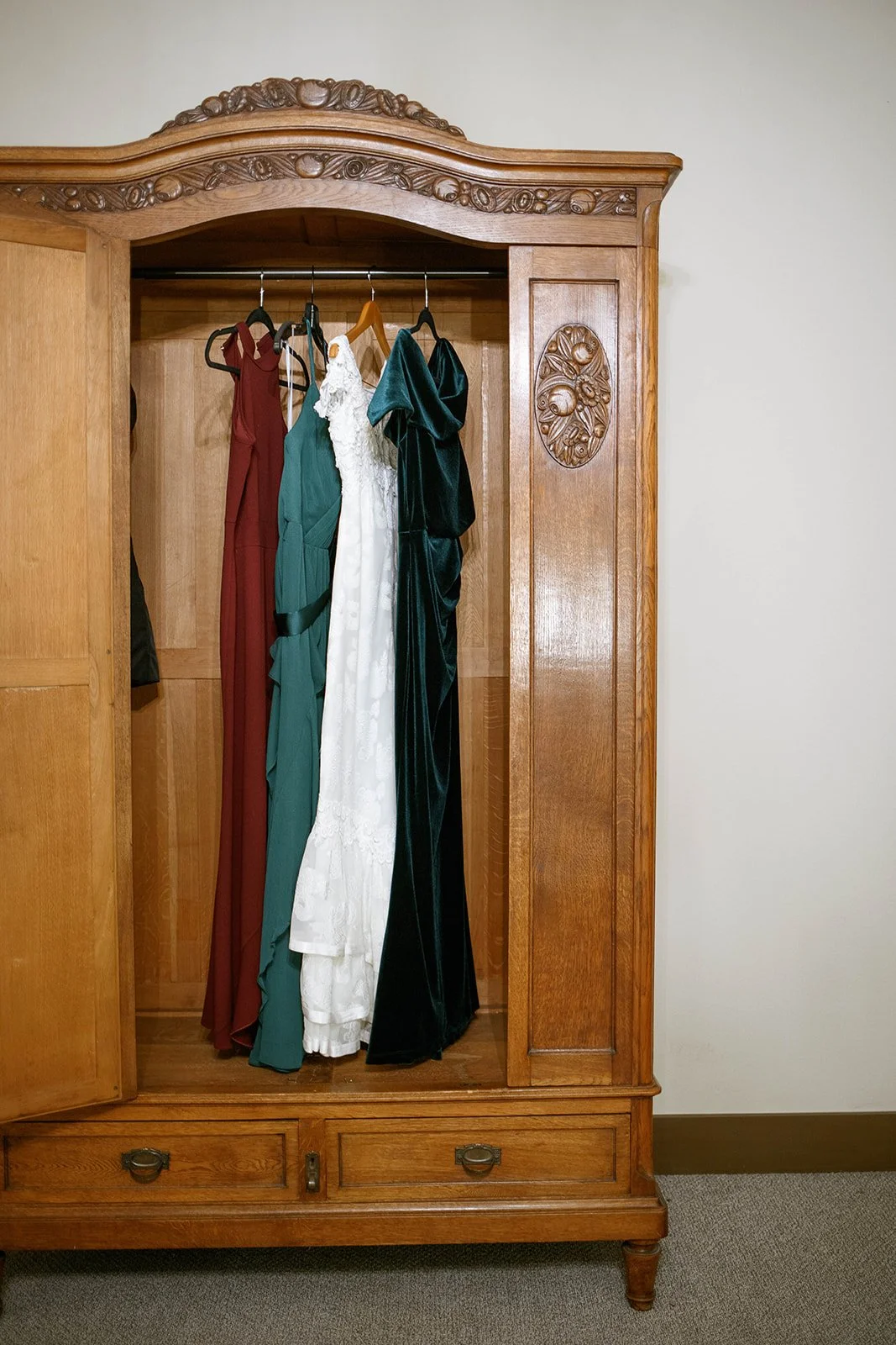 White bridal gown and jewel-tone bridesmaid dresses hanging inside a wooden wardrobe.