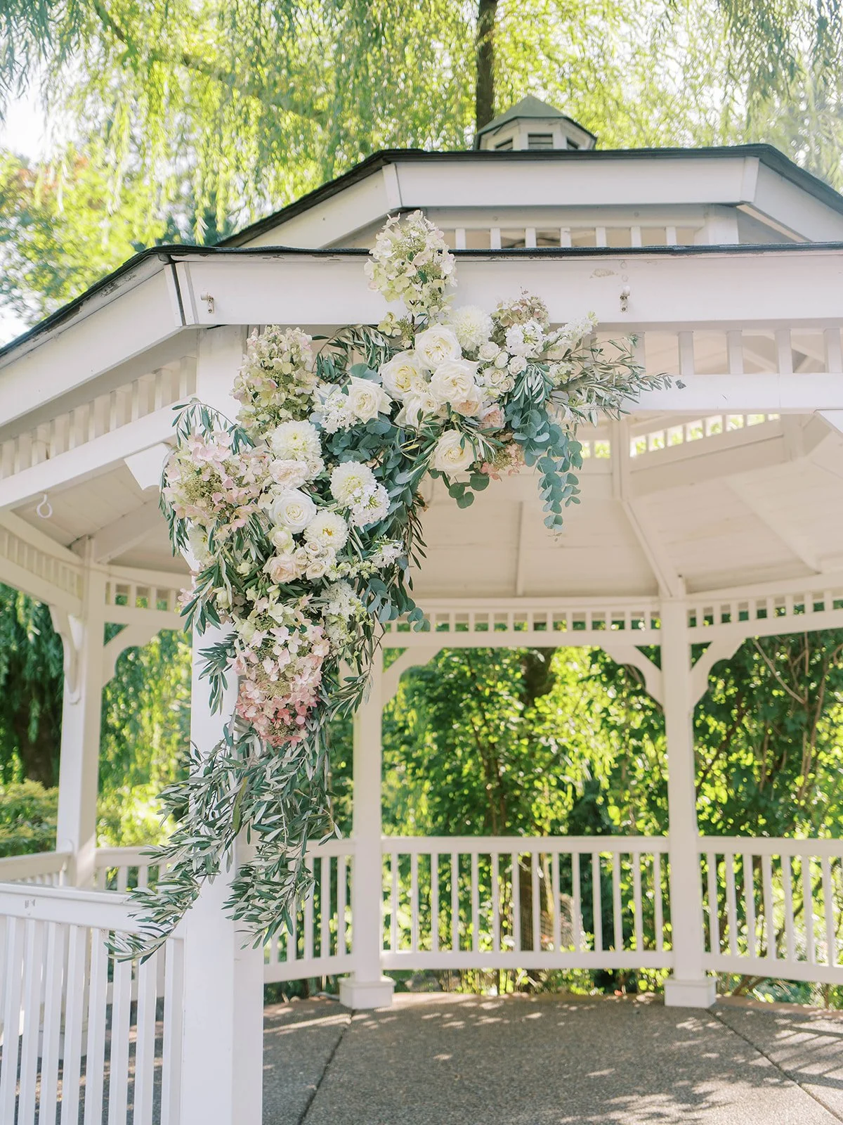 Elegant floral installation of white roses, blush hydrangeas, and olive branches on wedding gazebo.