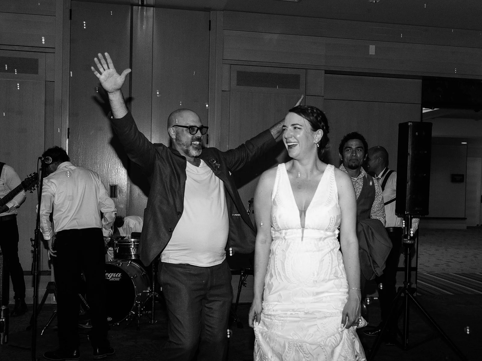 Black and white photo of the groom raising his arm in celebration while the bride laughs beside him near the band