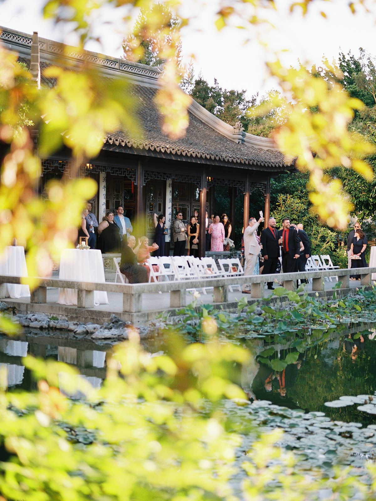 Wedding guests fill the ceremony terrace beside the lake at golden hour, viewed through soft bokeh foliage, white folding chairs arranged in rows before the pavilion.