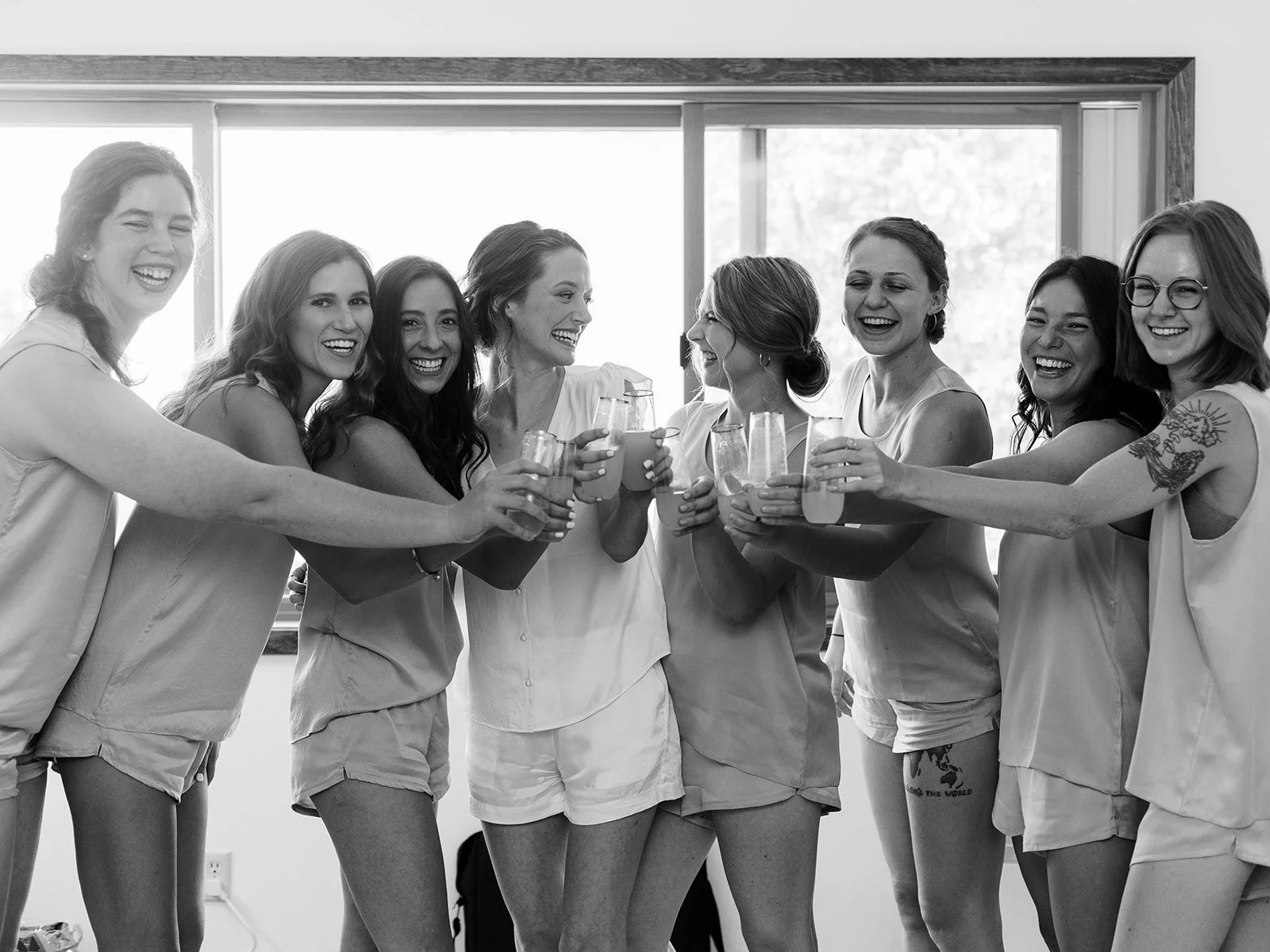 Bridal party in matching outfits clinking glasses and laughing indoors during wedding preparations.