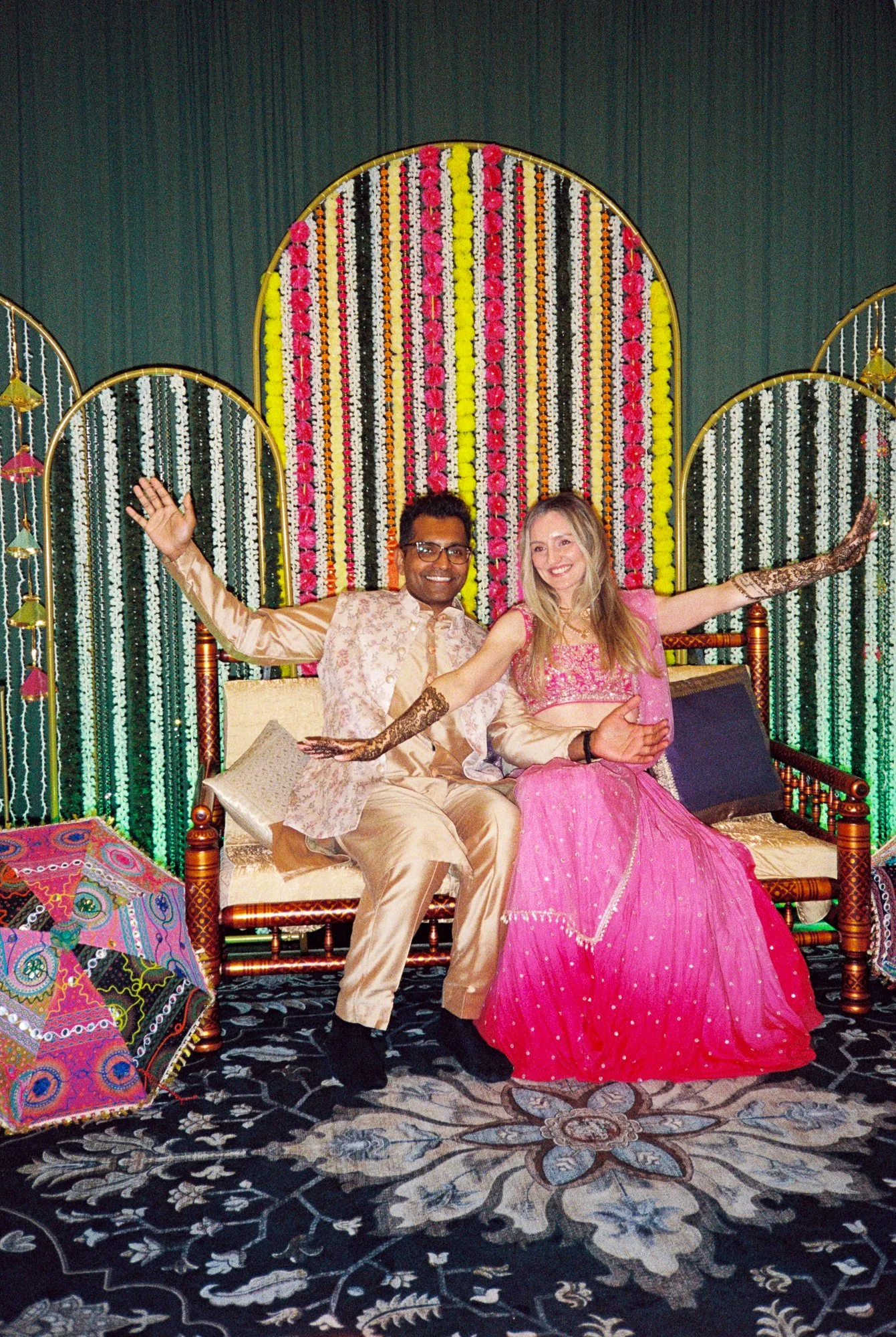 Alacia and Rohit seated together in front of a colorful floral Mehndi backdrop with arms outstretched.