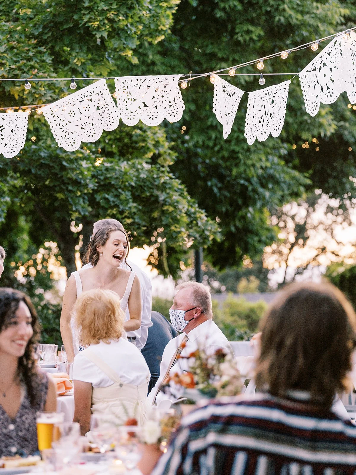 Bride laughing while speaking with guests at outdoor wedding reception under string lights and banners