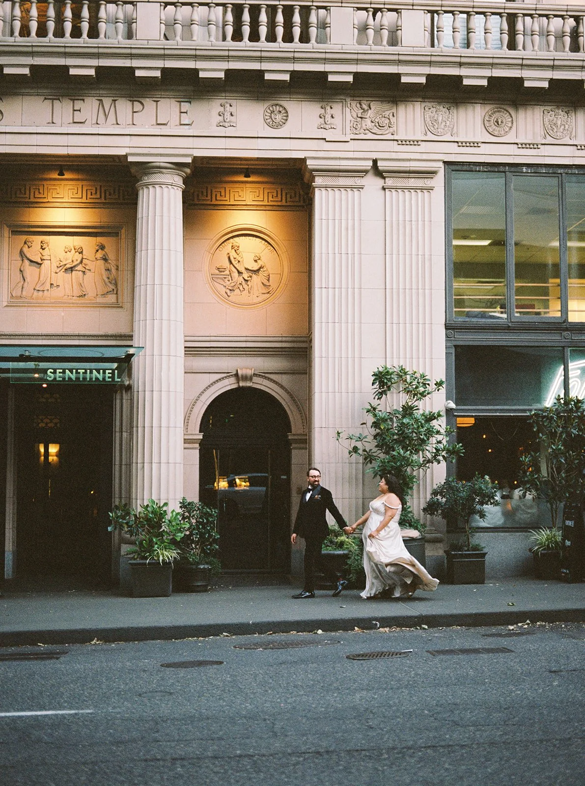 Couple holding hands while walking past ornate stone building with columns and carved facade.