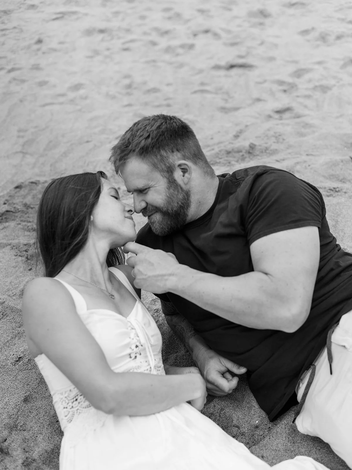 Black and white close-up of couple lying on sand, leaning in for a kiss.