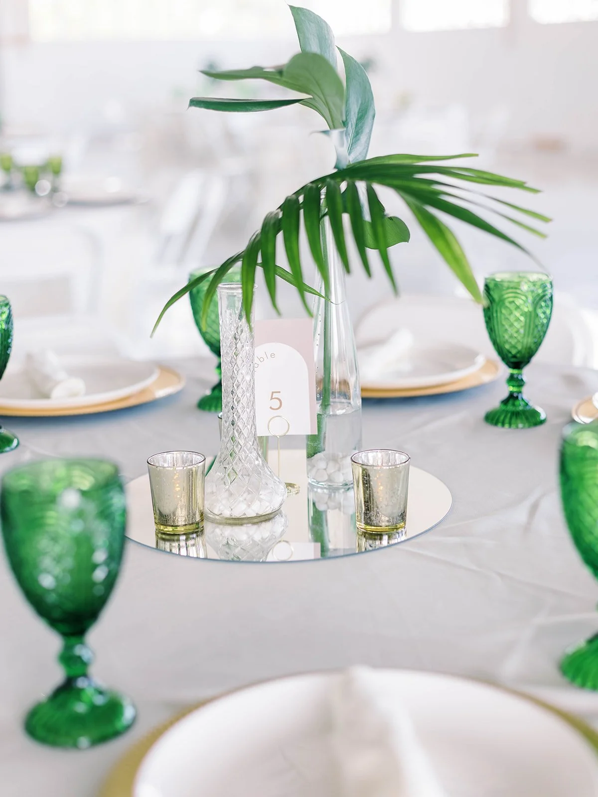 Close-up of a tropical wedding reception centerpiece featuring monstera and palm leaves in glass vases on a mirror base, surrounded by green goblets and a table number card
