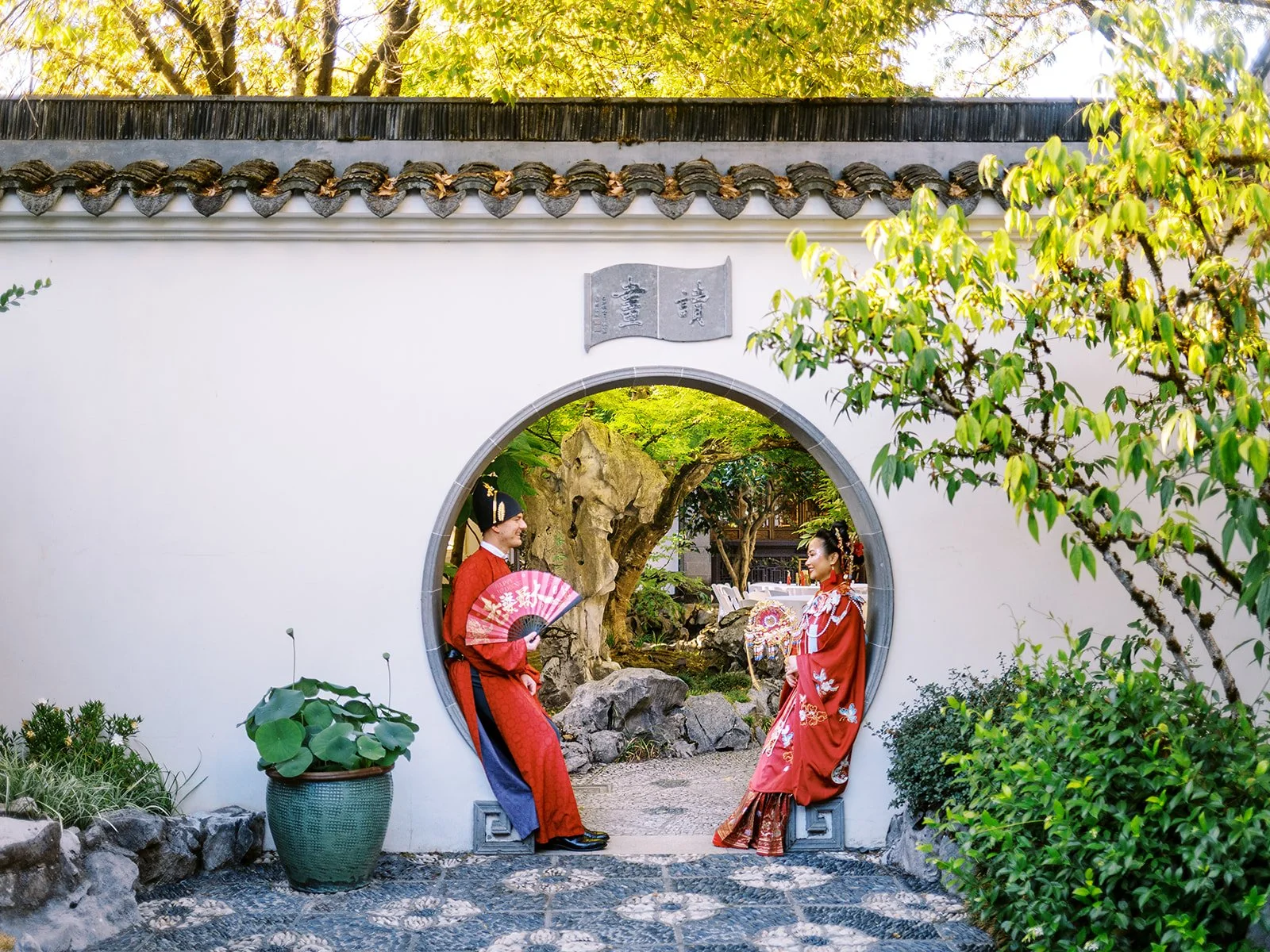 Wide shot of bride and groom sitting on opposite sides of a circular moon gate, facing each other in red traditional wedding attire with scholar's rocks and trees visible through the opening