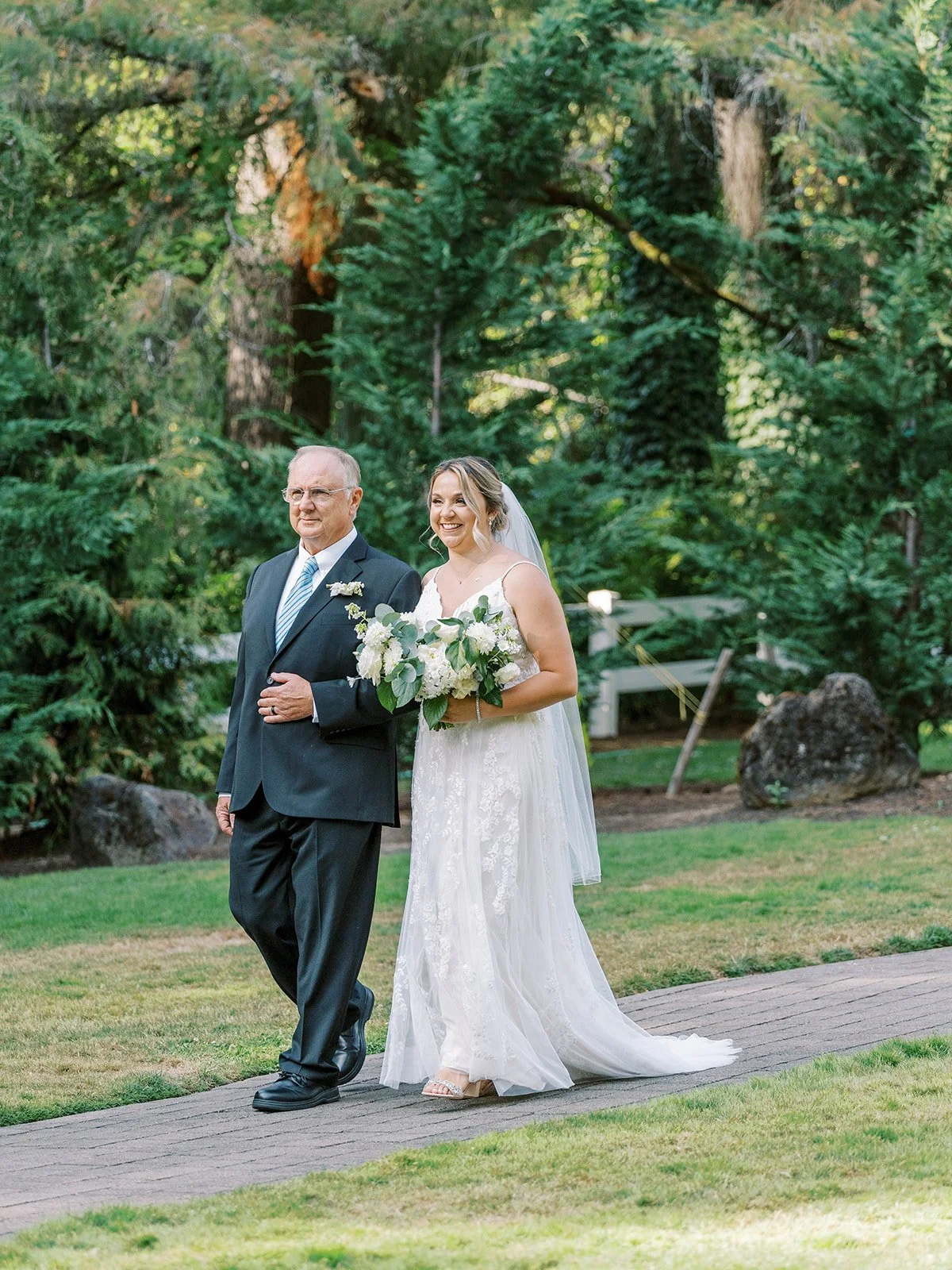 Bride and father smiling during outdoor wedding processional through lush garden setting.