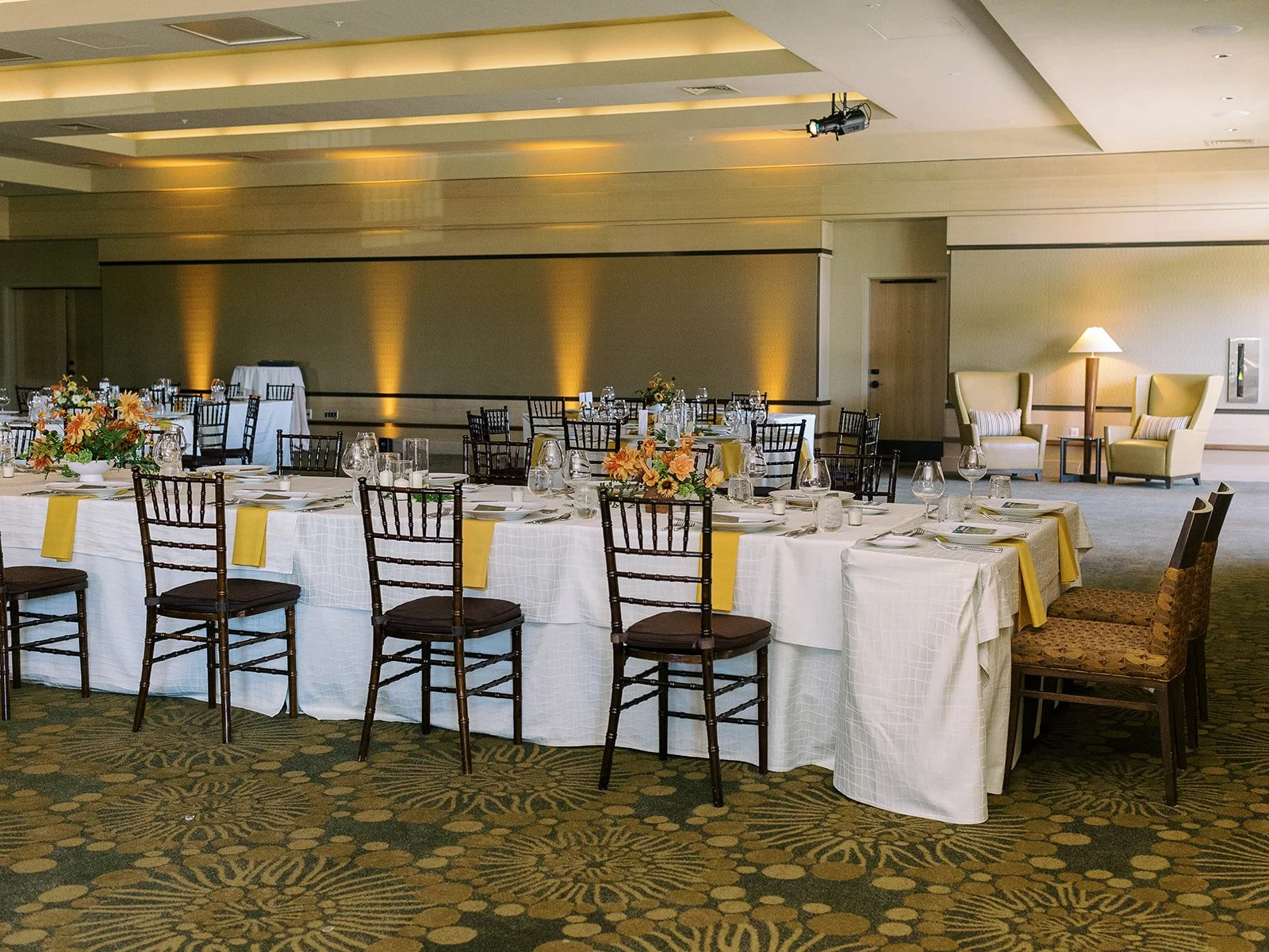 Wide shot of the wedding reception ballroom featuring a long rectangular head table set with white linens, yellow napkins, and orange floral centerpieces, with amber uplighting along the walls