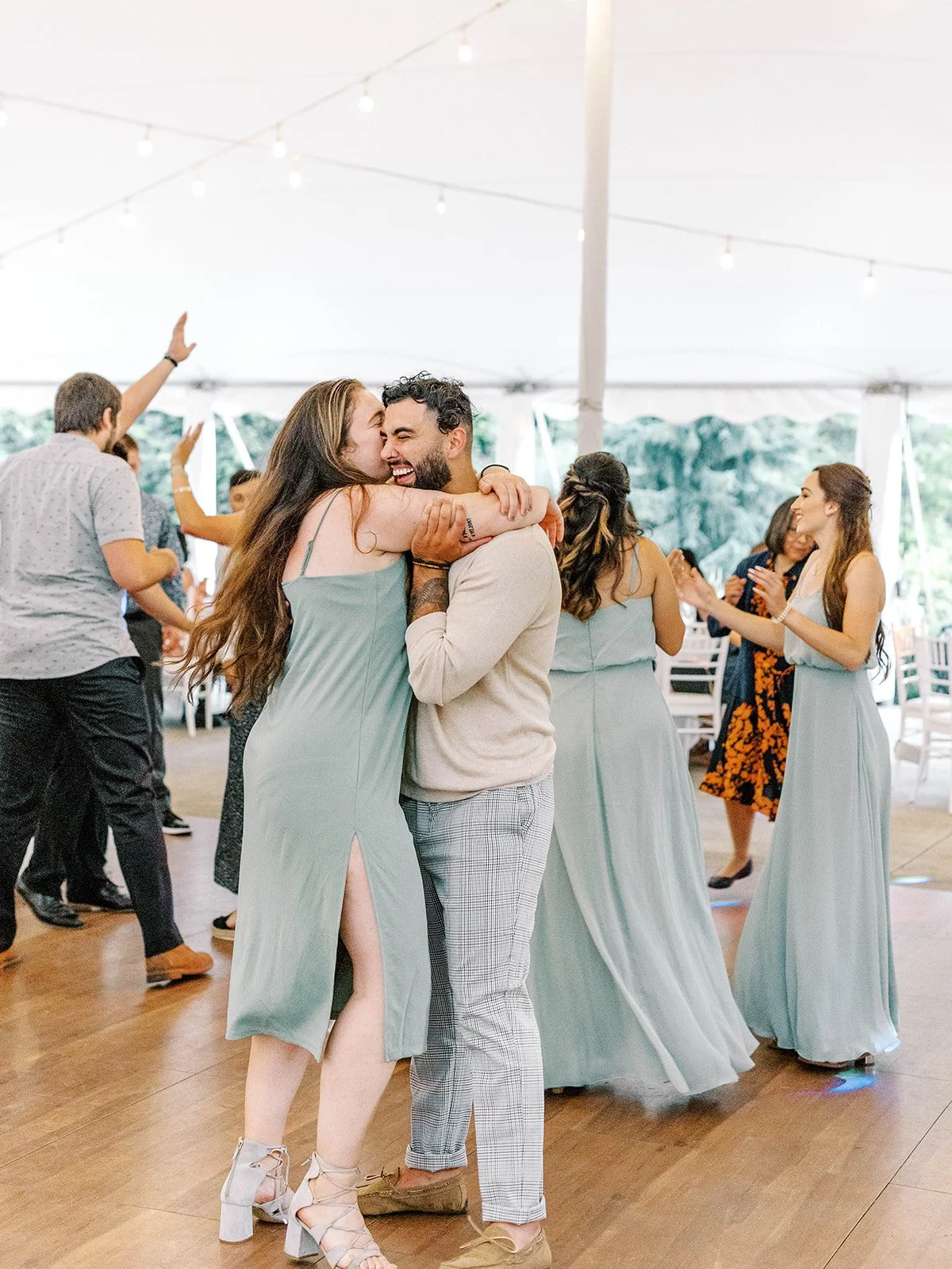 Wedding guests dance under a reception tent, including bridesmaids in sage green dresses celebrating on the dance floor.