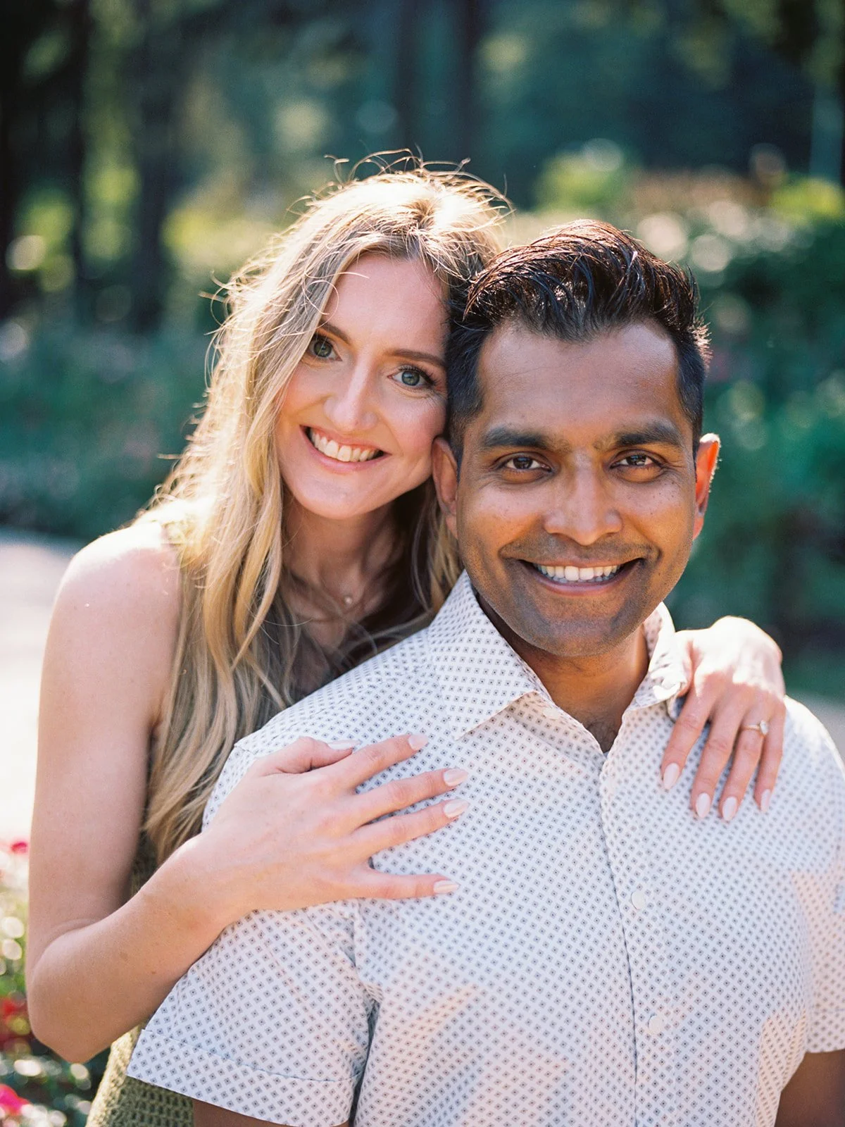 Close-up portrait of a smiling couple cheek-to-cheek in a sunlit garden, she draped over his shoulder from behind.