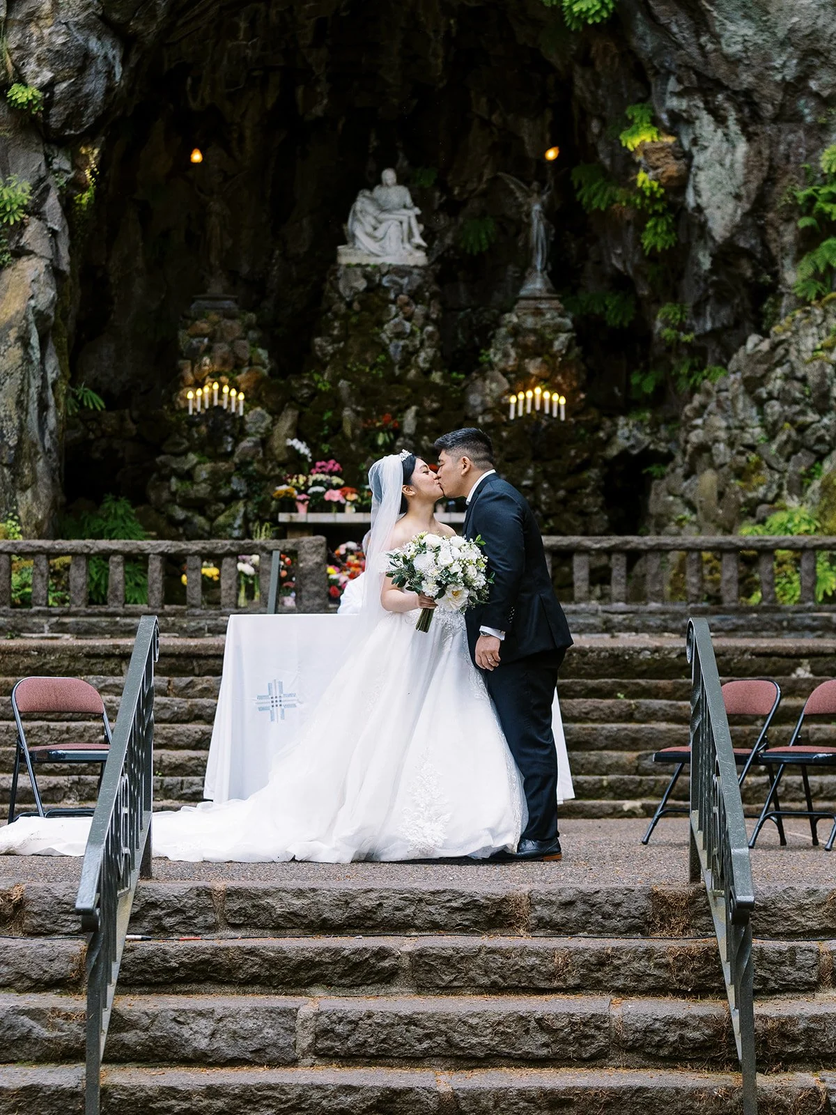 Bride and groom sharing their first kiss at The Grotto's cave altar, the Pietà sculpture and glowing candelabras visible in the rock grotto behind them