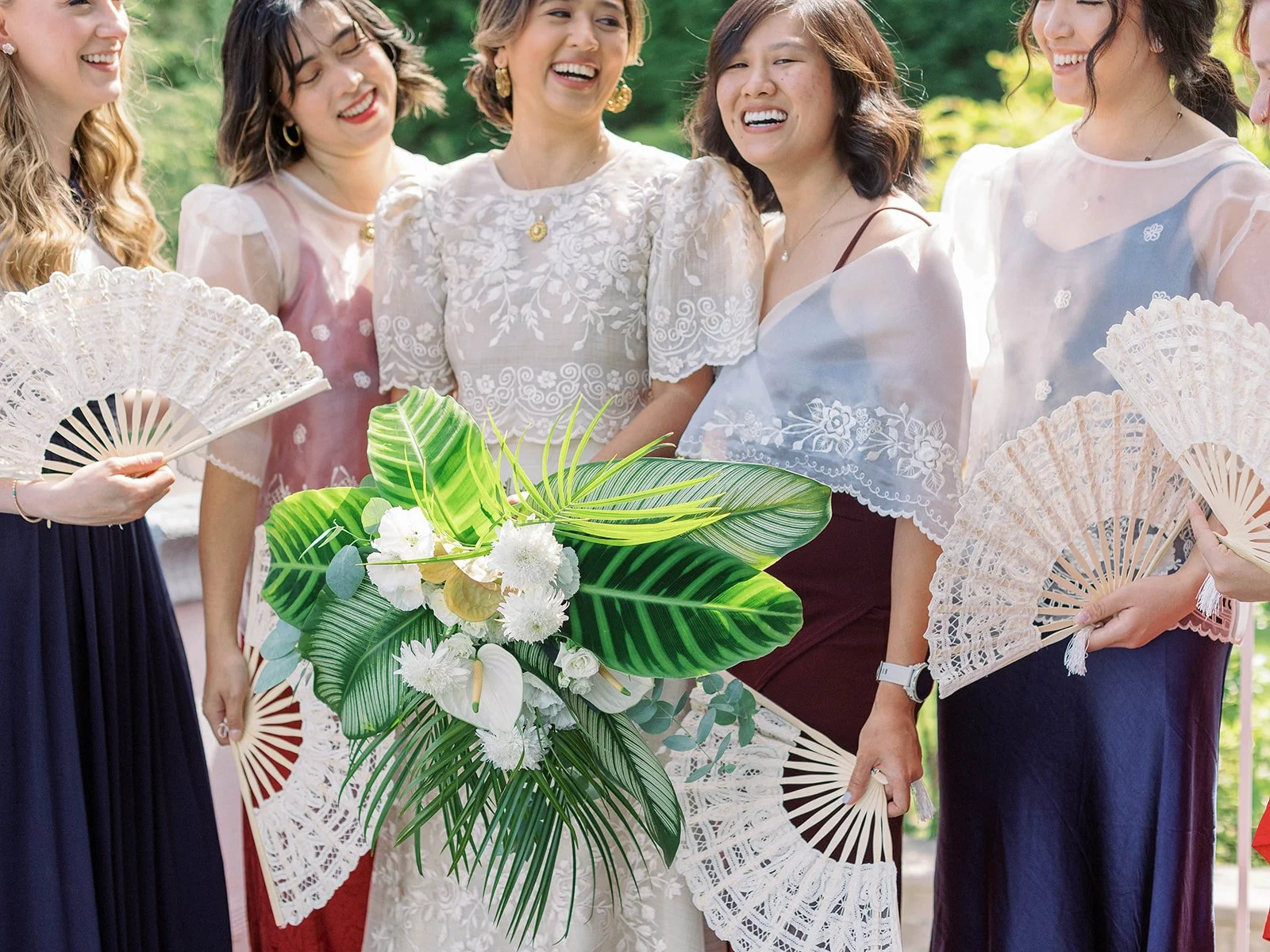 Bride and bridesmaids laughing together outdoors, holding white lace fans, with a lush tropical bouquet in the foreground
