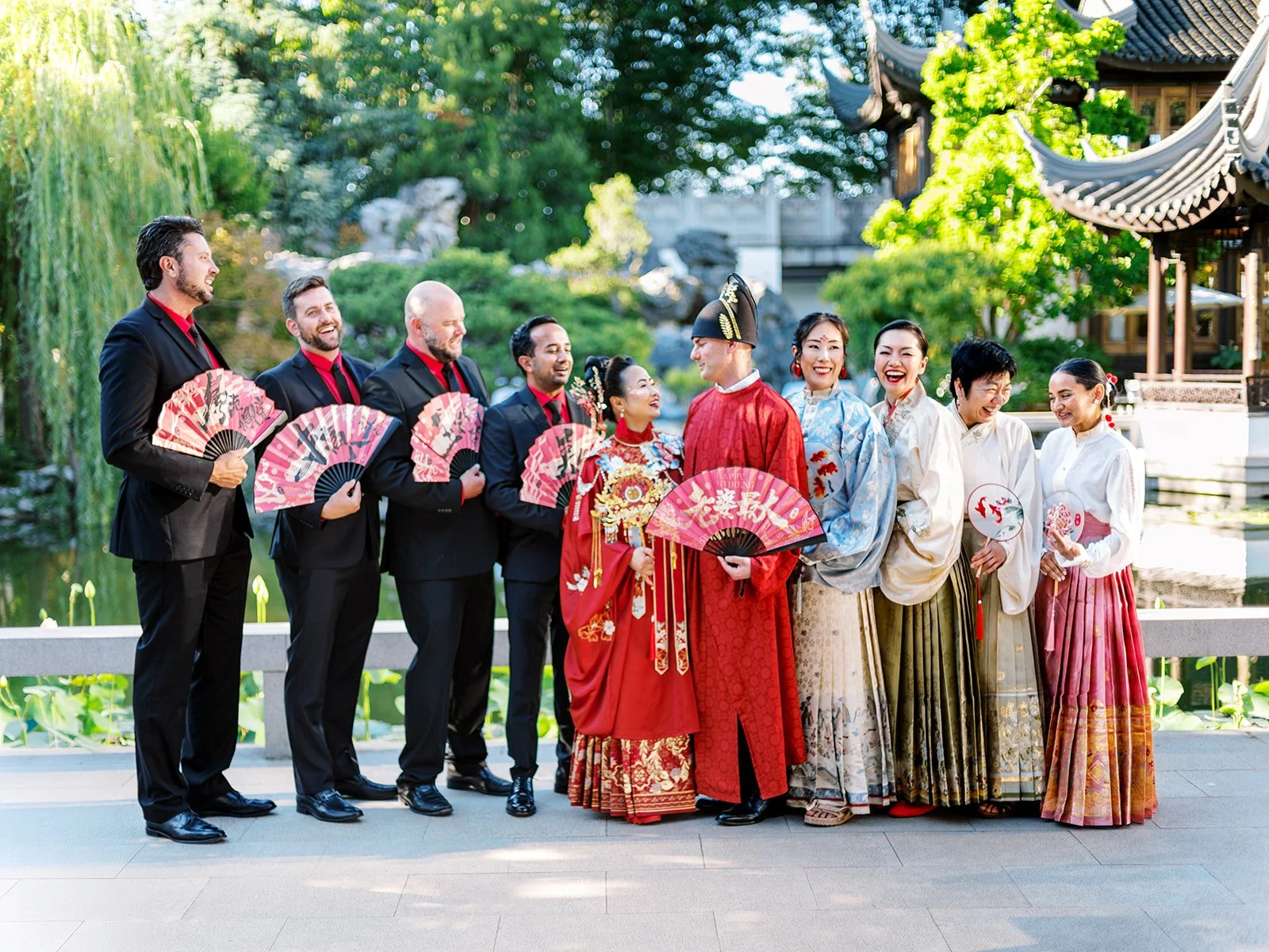 Full wedding party at Lan Su Chinese Garden's koi pond, groomsmen in black suits with red shirts holding red fans alongside the wedding party in hanfu