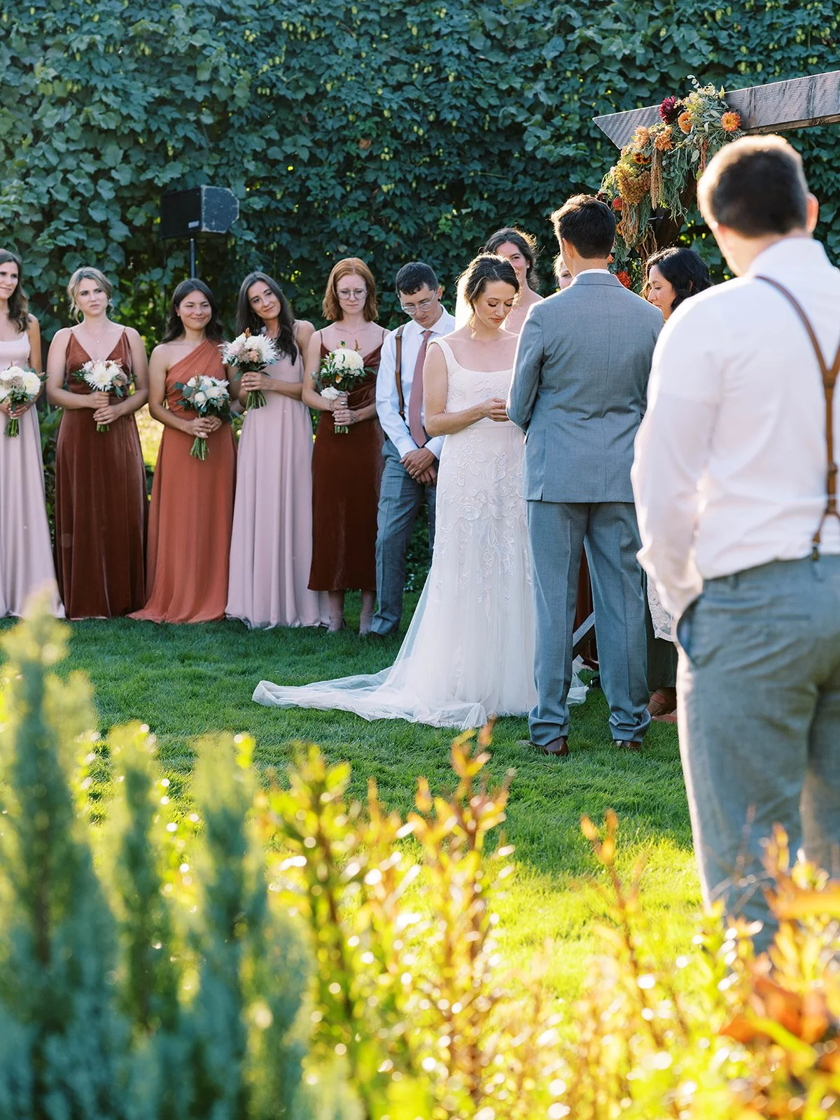 Bride and groom standing together during an outdoor garden wedding ceremony with their wedding party behind them.
