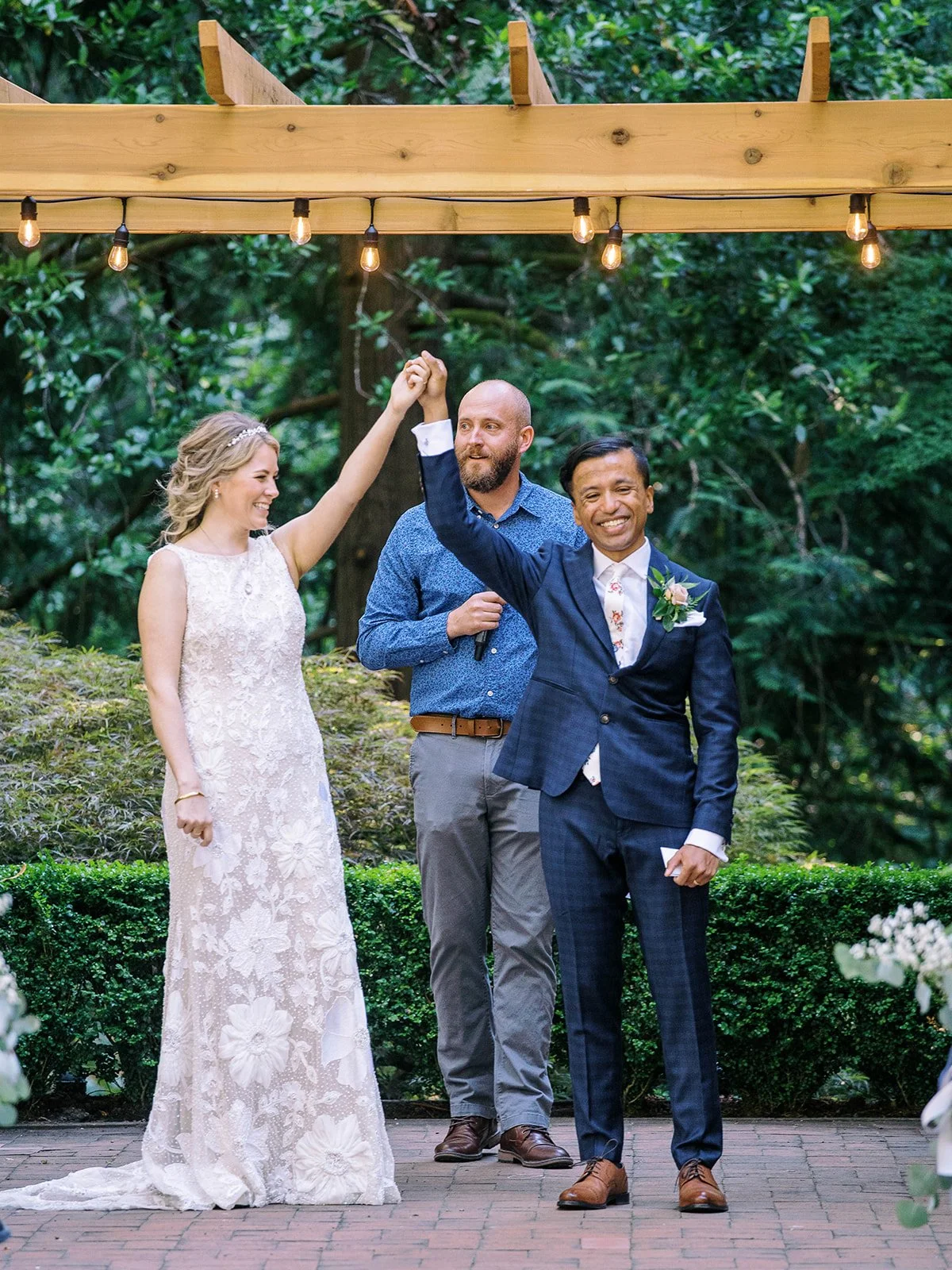 Newlyweds raise their joined hands after the ceremony beneath string lights and greenery.