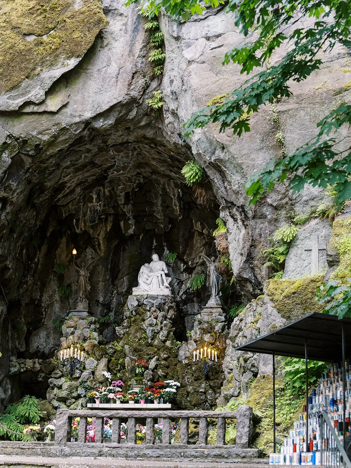 The cave grotto shrine at The Grotto Portland, featuring a Pietà sculpture, candles, and colorful floral offerings set into a mossy basalt rock face