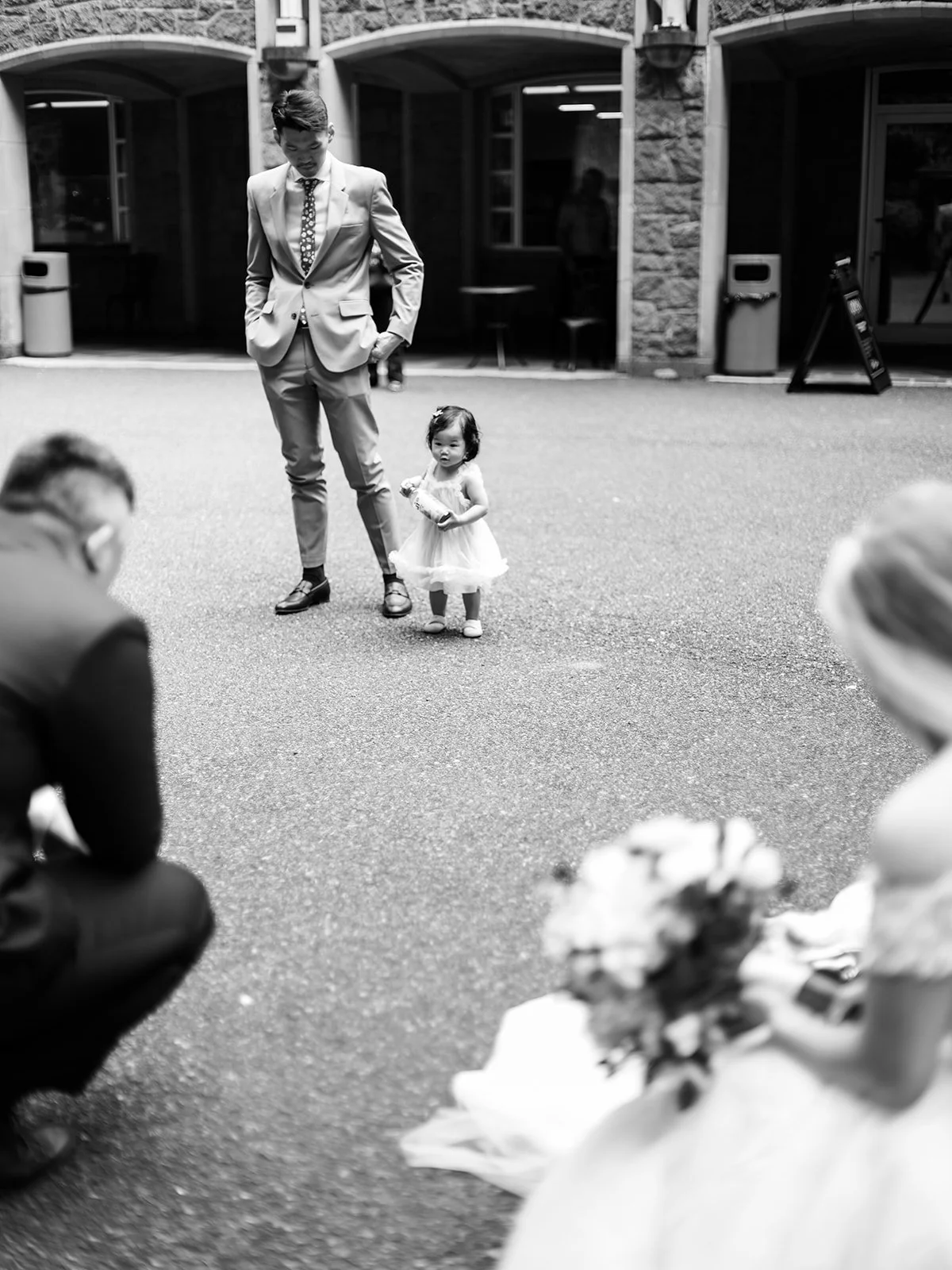Black and white candid of a tiny flower girl in a white tulle dress standing beside a man in a tan suit in The Grotto's courtyard, the bride blurred in the foreground
