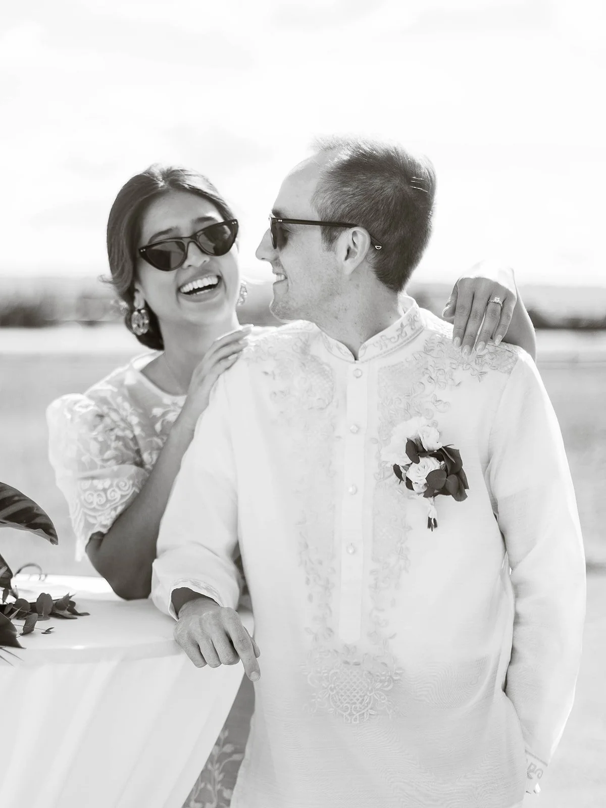 Black and white portrait of bride and groom laughing together in sunglasses while leaning on a cocktail table at the airfield
