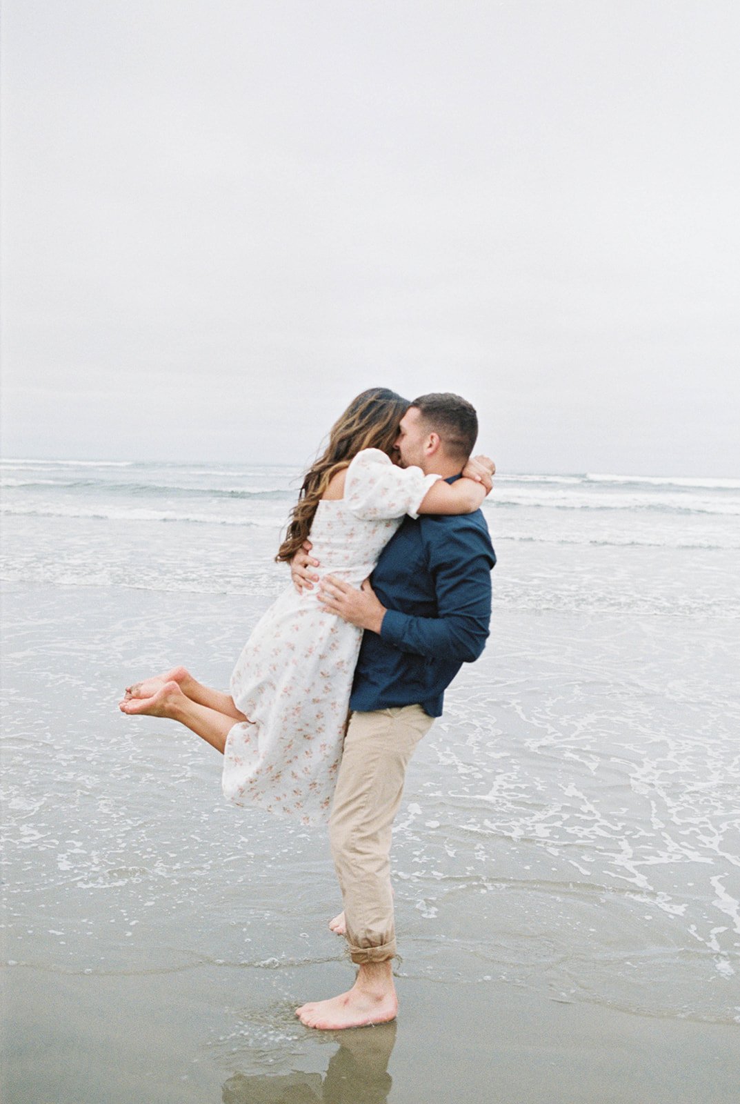 Man lifting woman while kissing in shallow ocean water during beach engagement session