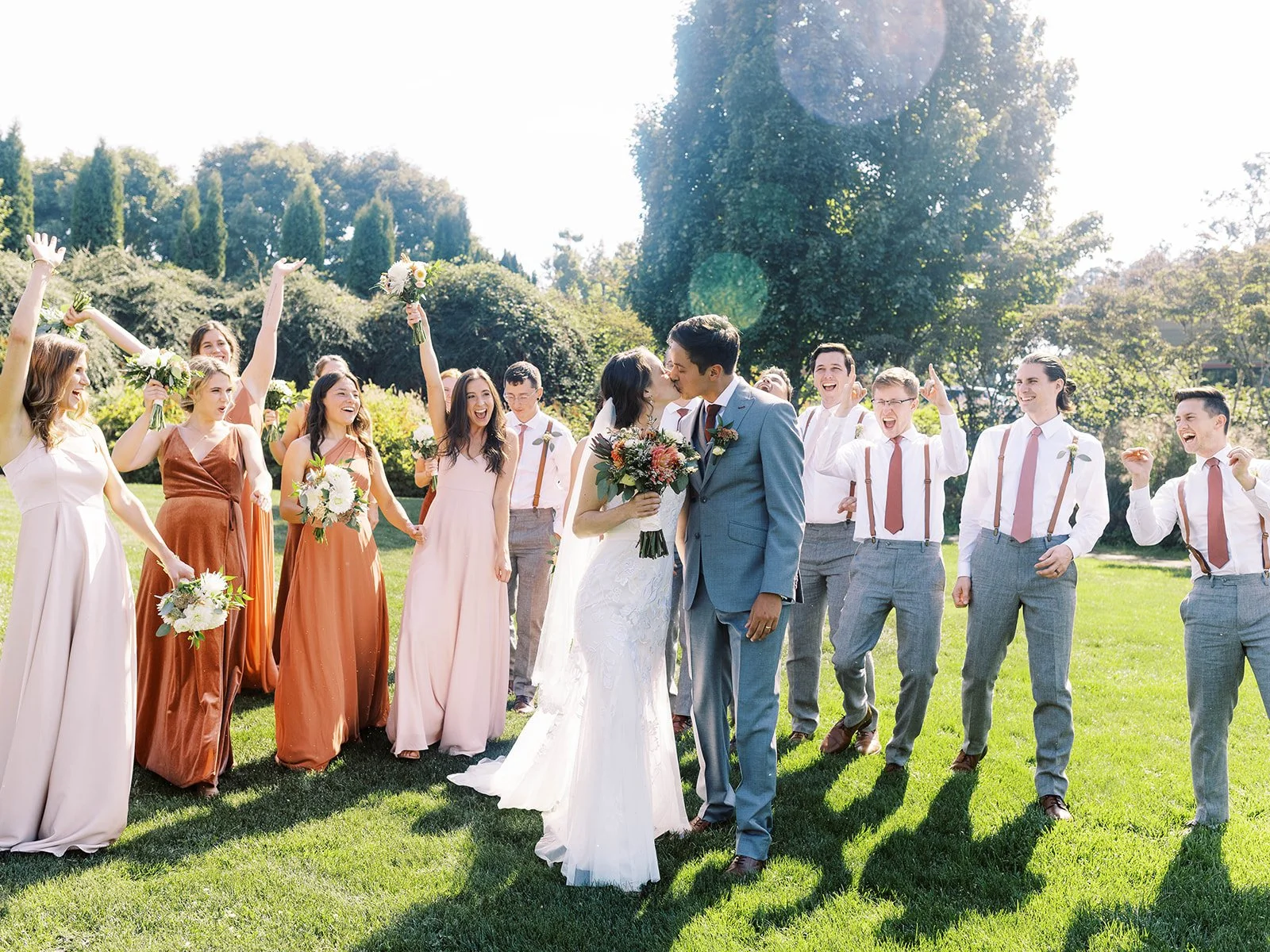 Bride and groom kissing on the lawn while their wedding party celebrates around them.