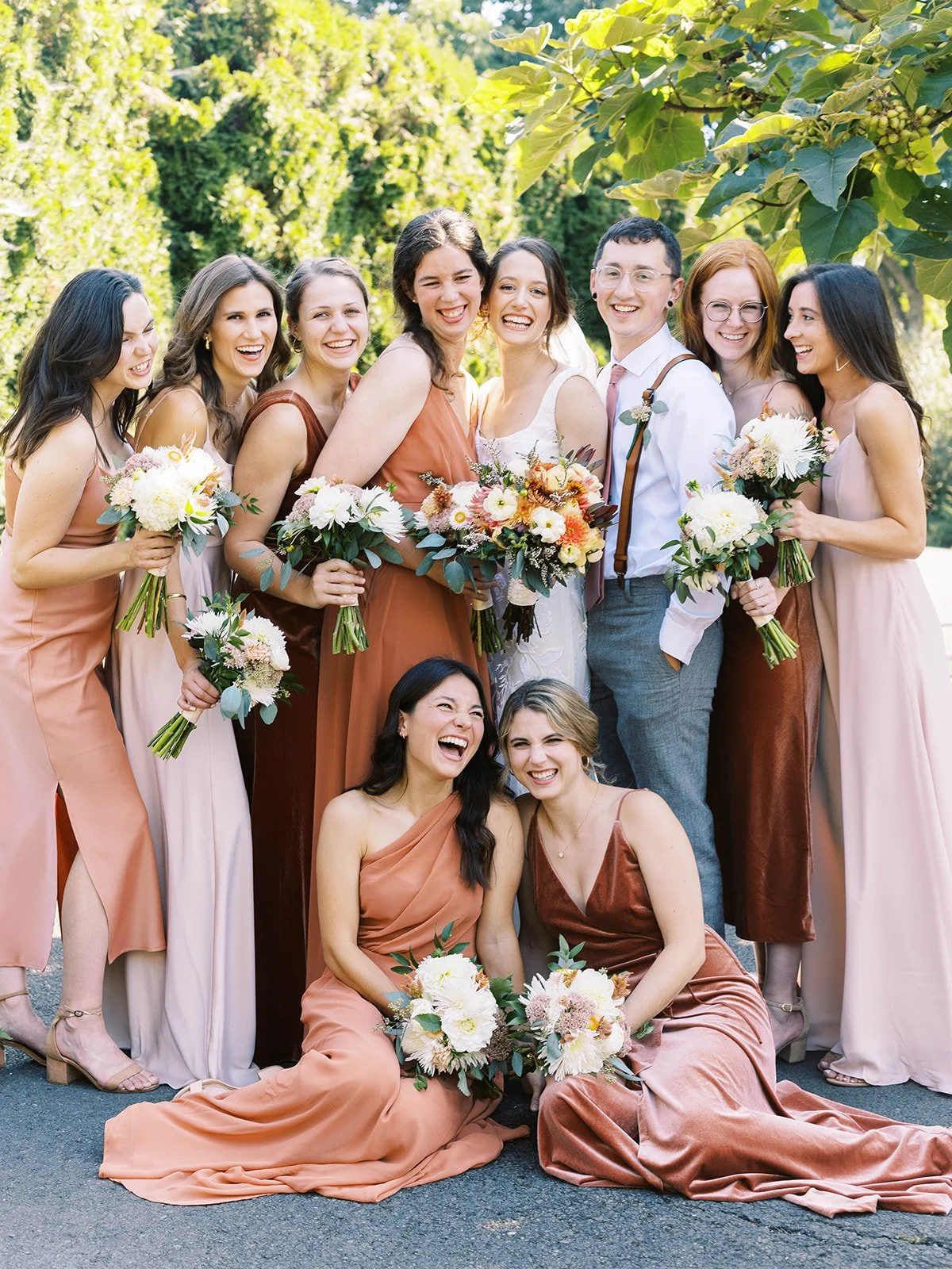 Wedding party posing together outdoors in blush, rust and gray attire while holding floral bouquets.