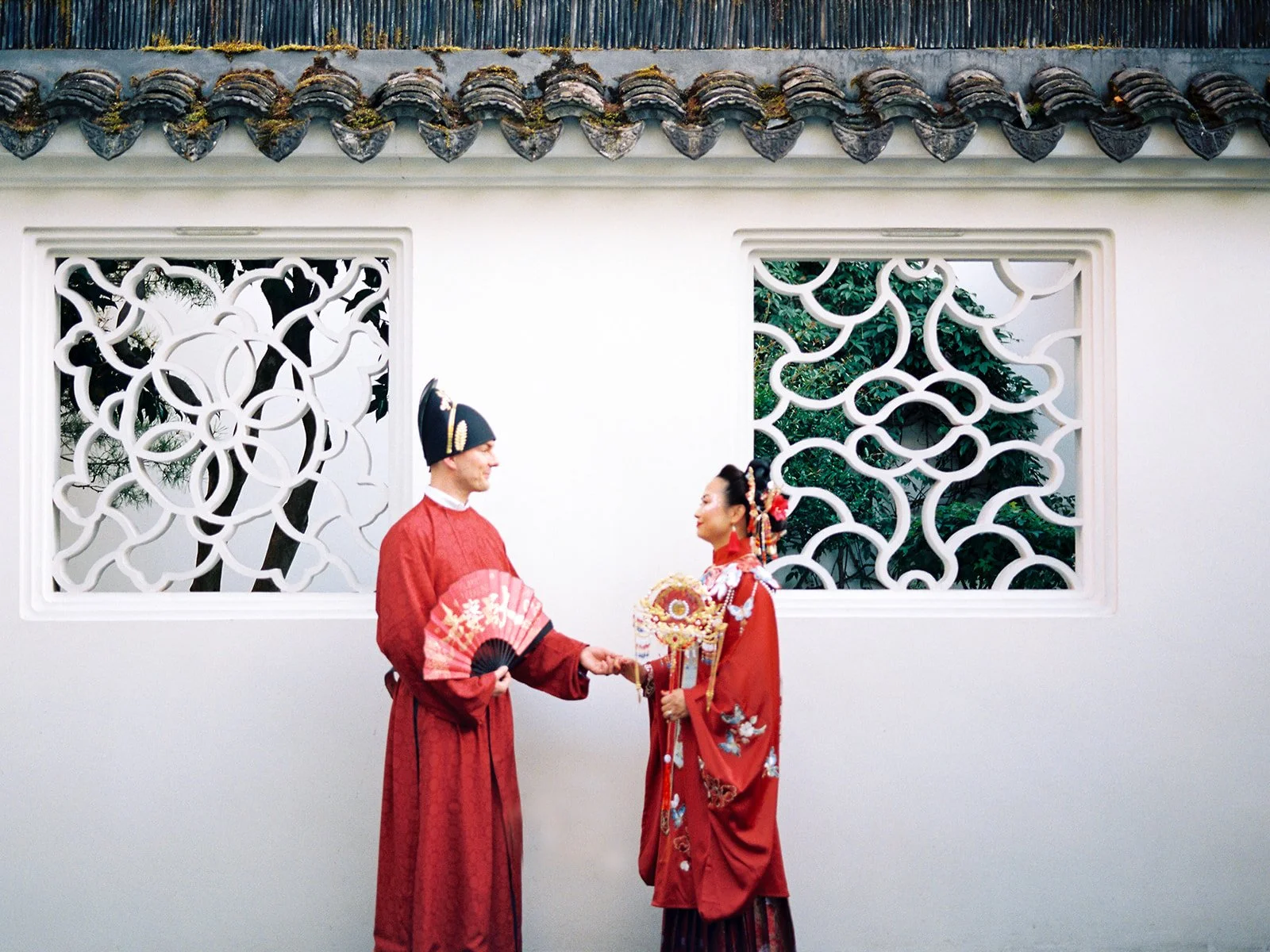 Bride and groom in matching red traditional Chinese wedding attire holding hands and facing each other in front of two ornate lattice windows beneath a tiled roofline at Lan Su Chinese Garden