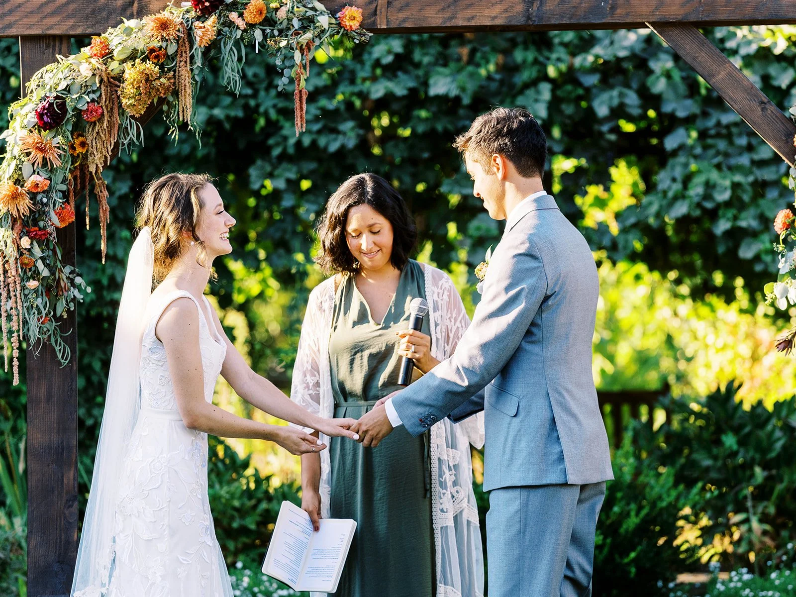 Couple holding hands during wedding ceremony with officiant under floral arch in garden