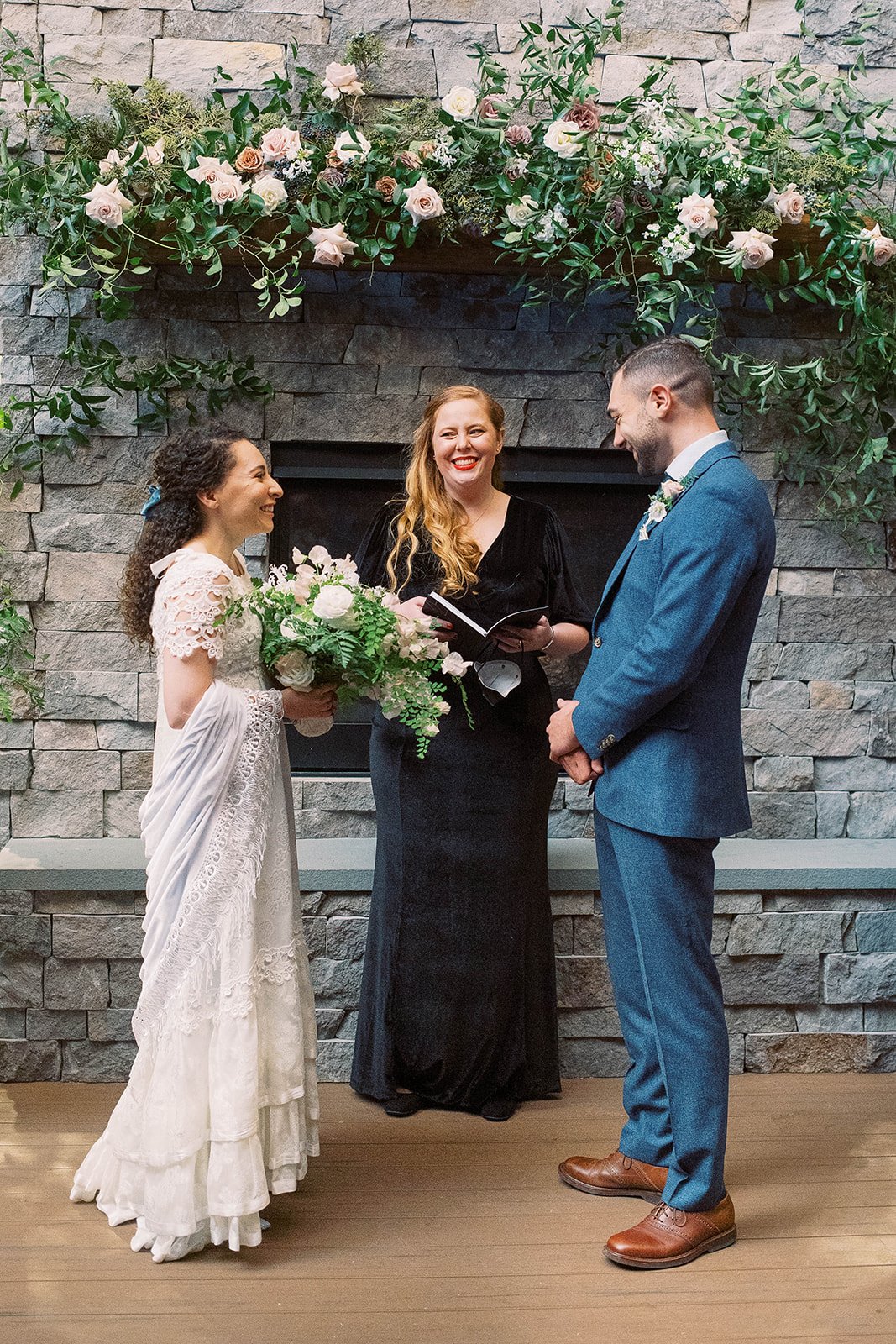 Bride and groom exchange vows with their officiant beneath lush flowers and greenery on a stone fireplace.