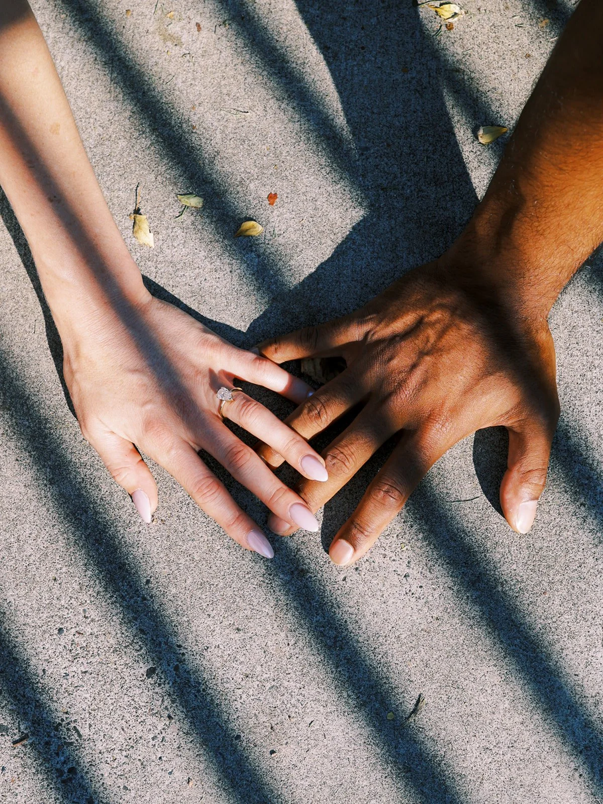 An overhead shot of two hands resting together on stone, the engagement ring catching the light amid striped shadows.
